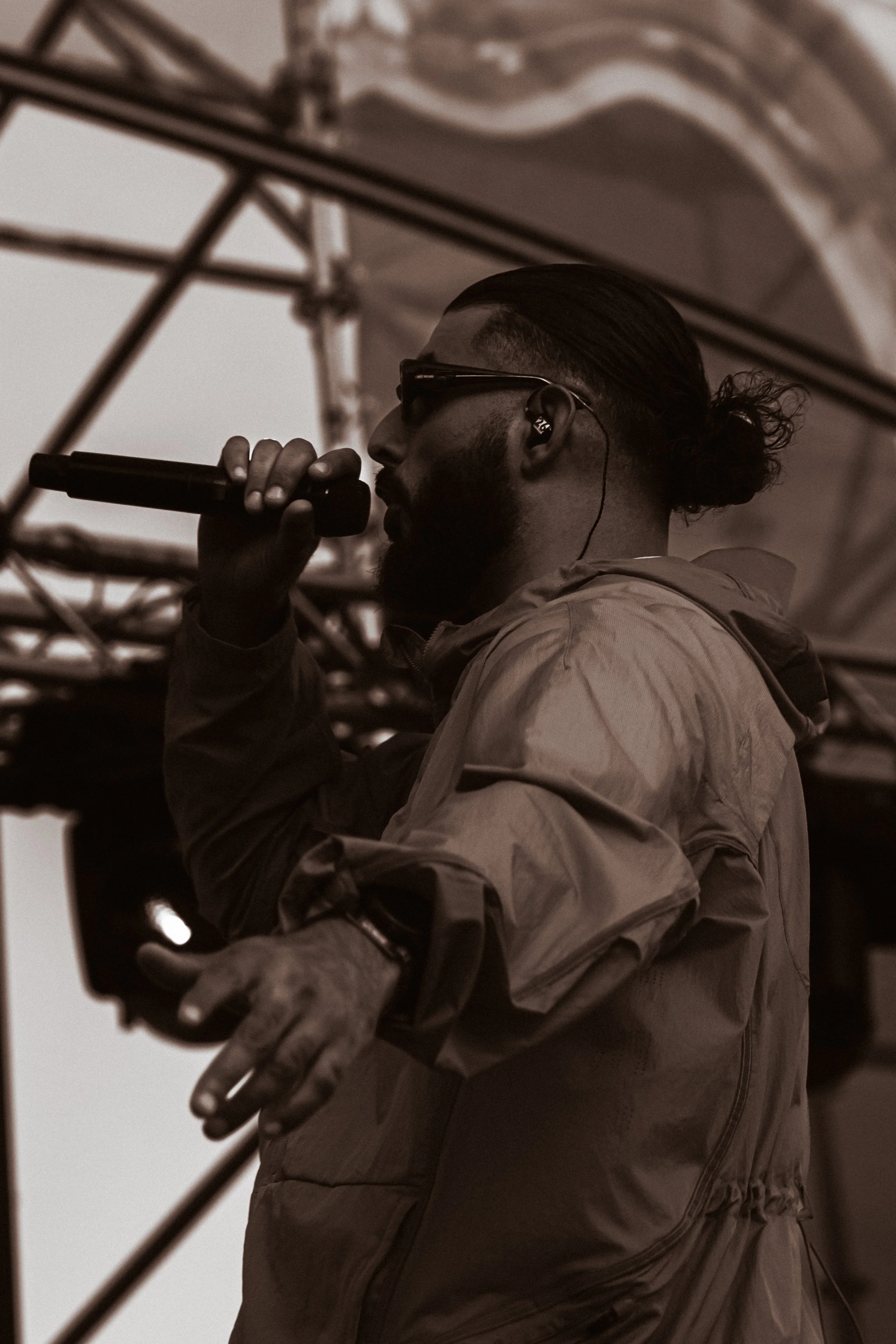 Un homme avec une barbe, portant des lunettes de soleil, écouteurs et un manteau, chante ou parle dans un microphone lors d'une performance en extérieur.