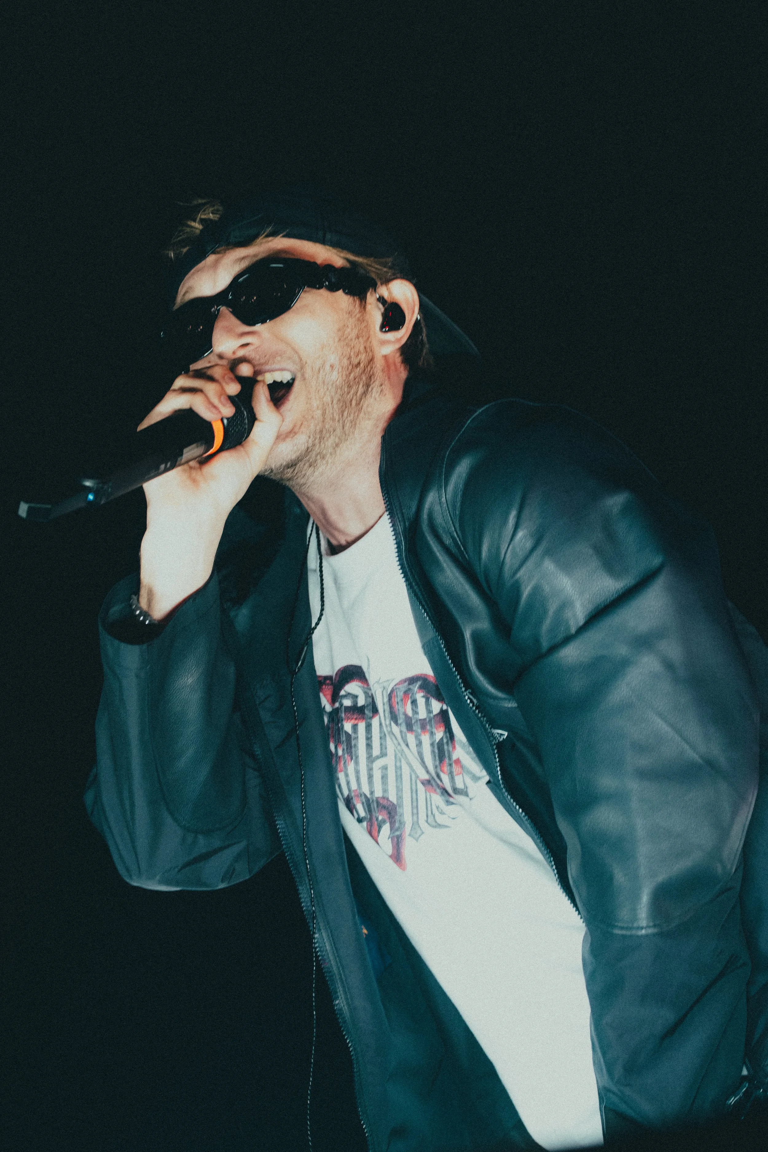 Un homme en veston en cuir noir portant des lunettes de soleil et un casque, chantant dans un micro lors d'une performance.