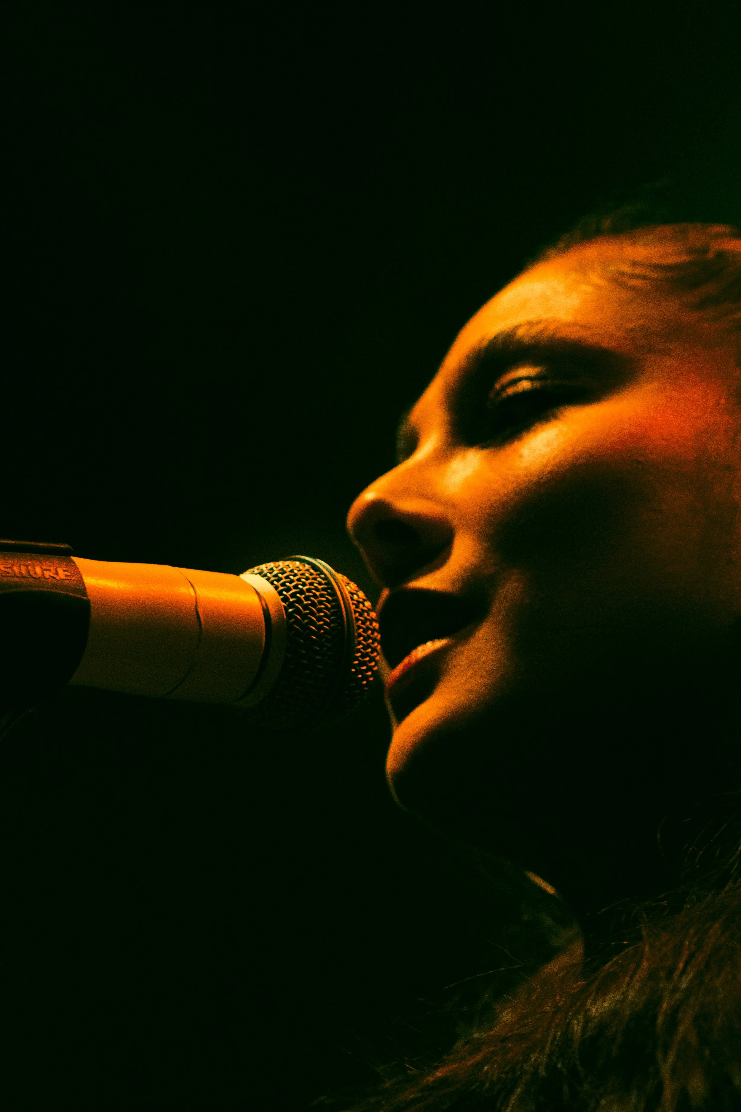Une femme chante ou parle dans un microphone, éclairée par une lumière chaude, sur un fond sombre.