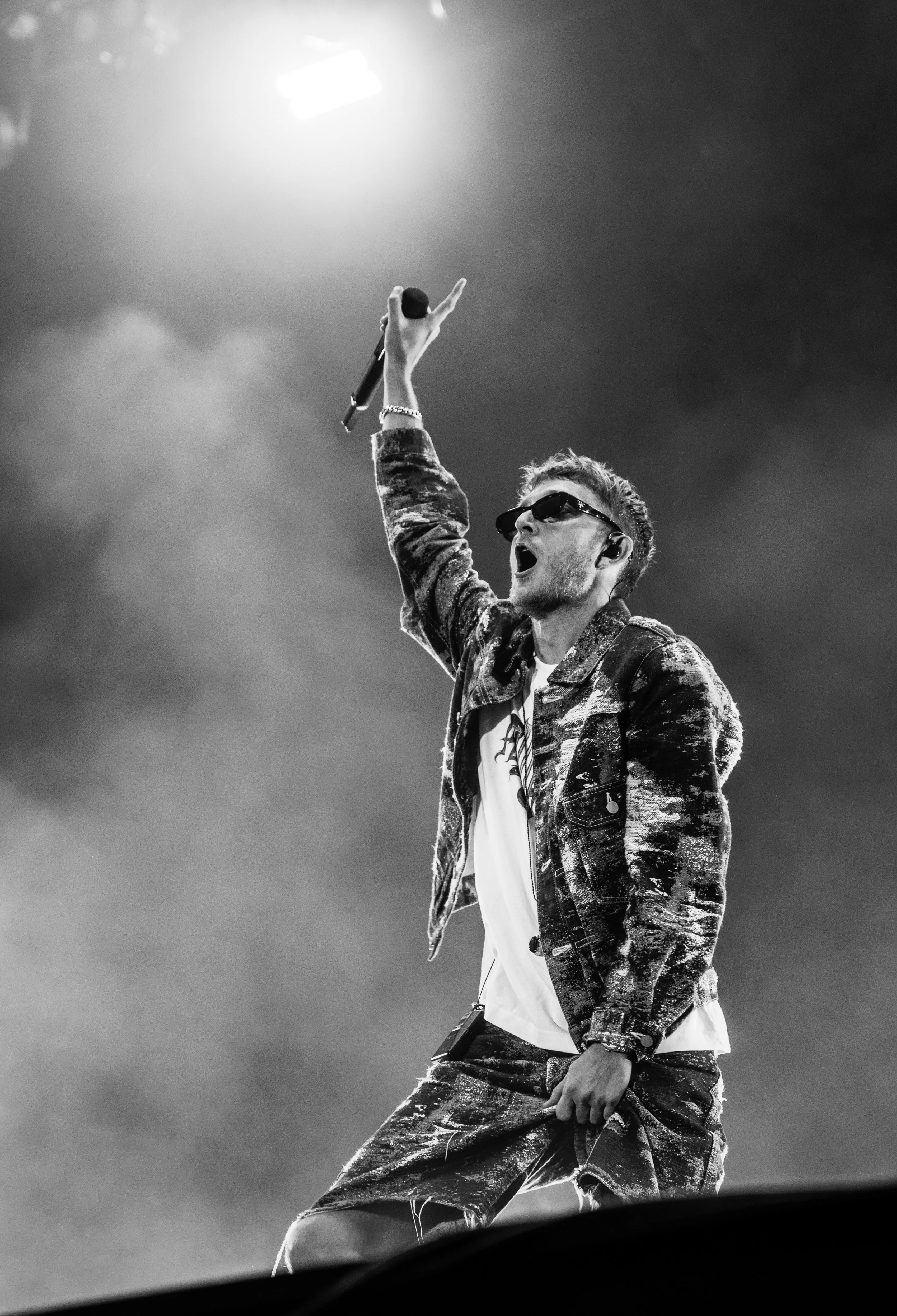 Un homme portant des lunettes de soleil, un jean et un sweat-shirt, tient un micro en l'air lors d'une performance sur scène en noir et blanc.