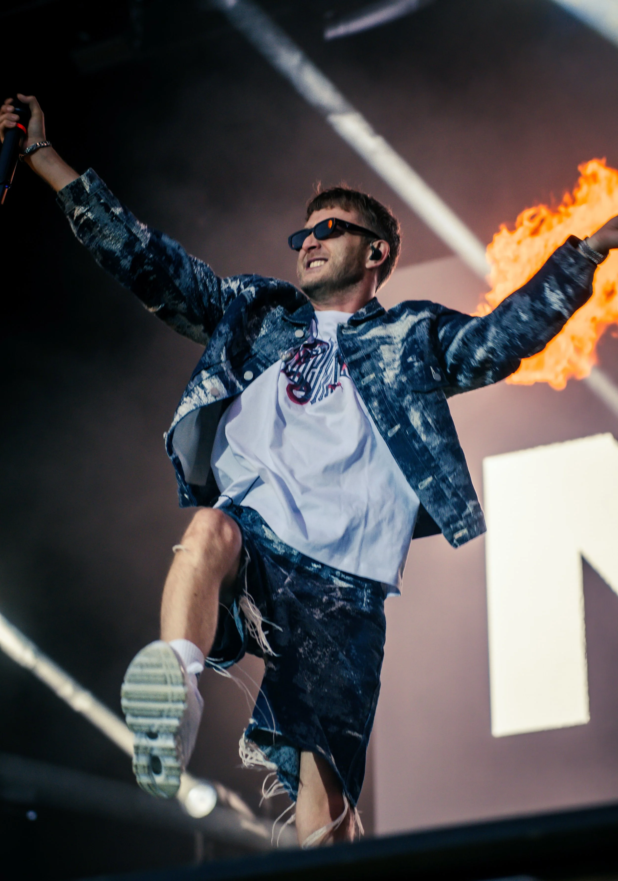 Un homme en tenue décontractée, portant des lunettes de soleil, chante ou danse sur scène avec des flammes en arrière-plan, lors d'un concert ou d'un spectacle.