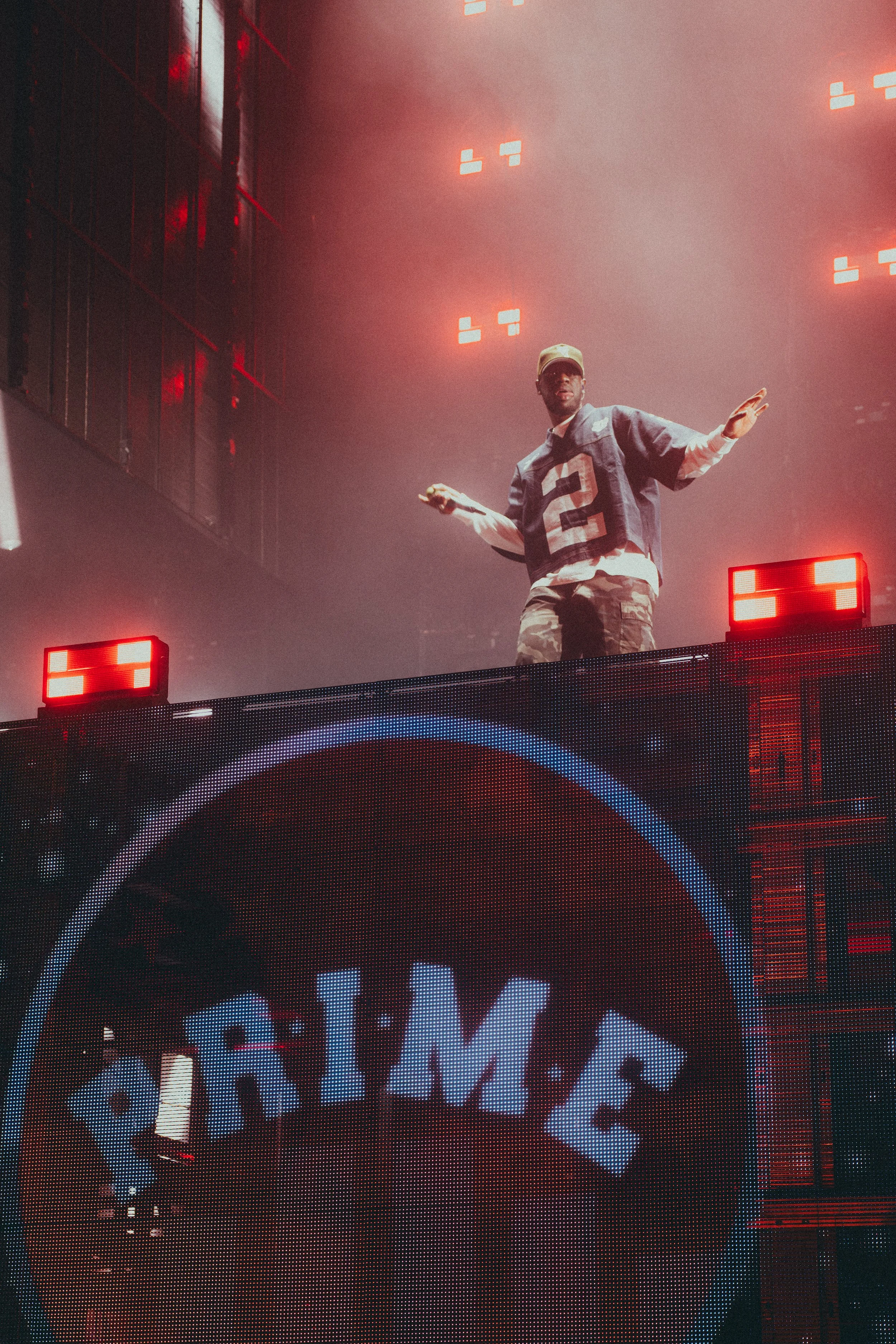 Un DJ en train de mixer sur une scène avec un écran LED affichant le nom « PRIME » et des lumières rouges, dans une ambiance de concert ou de fête.