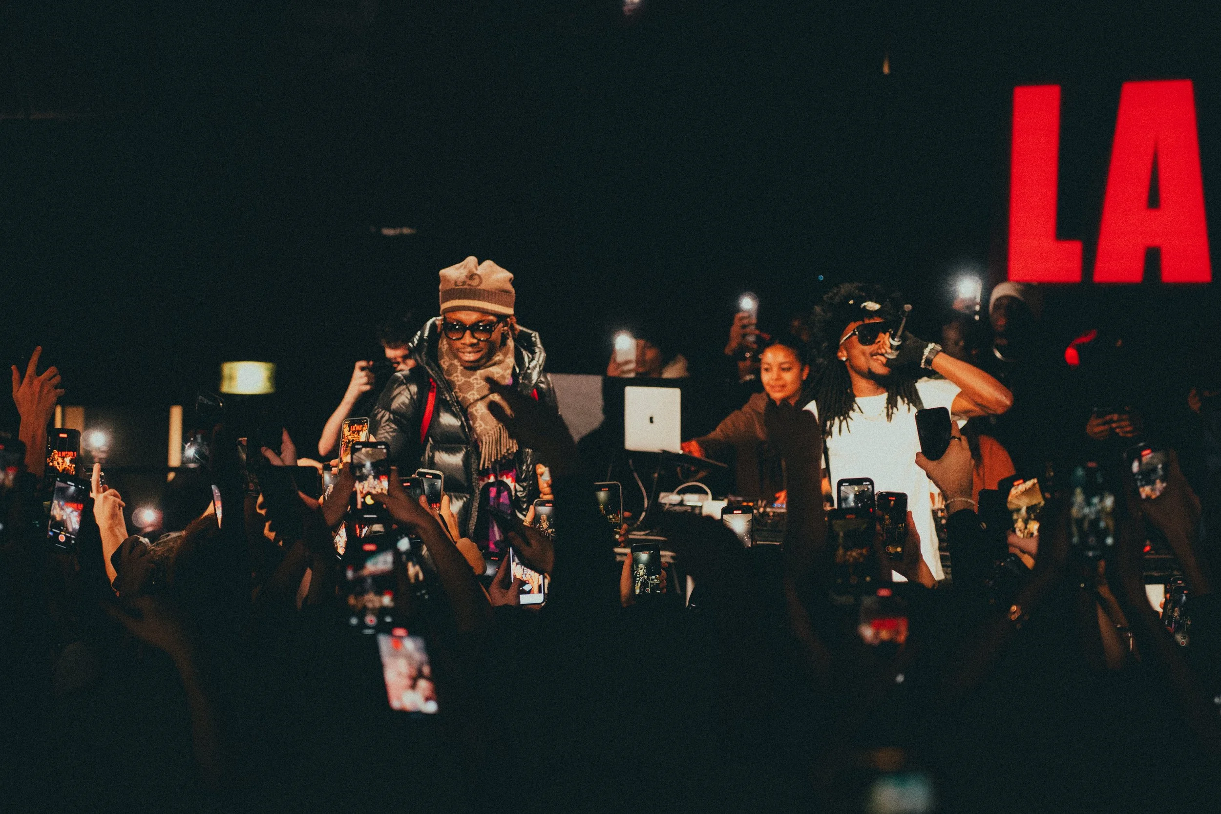 Deux artistes ou DJ sur scène lors d'un concert, entourés de nombreux spectateurs tenant des smartphones, avec un grand panneau lumineux rouge affichant 'LA' en arrière-plan.