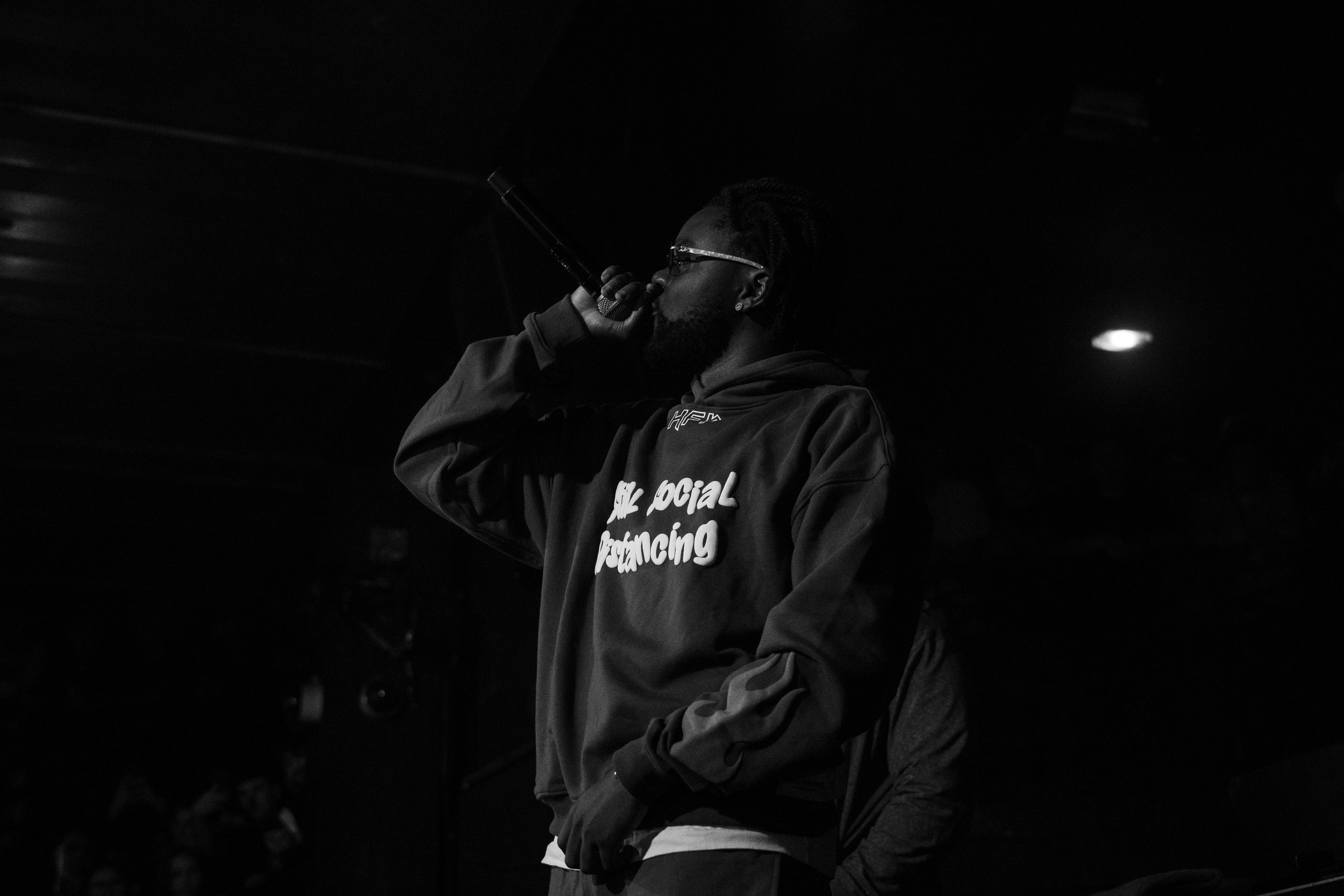 Un homme avec lunettes, portant un sweat à capuche, chante dans un micro sur scène, en noir et blanc.