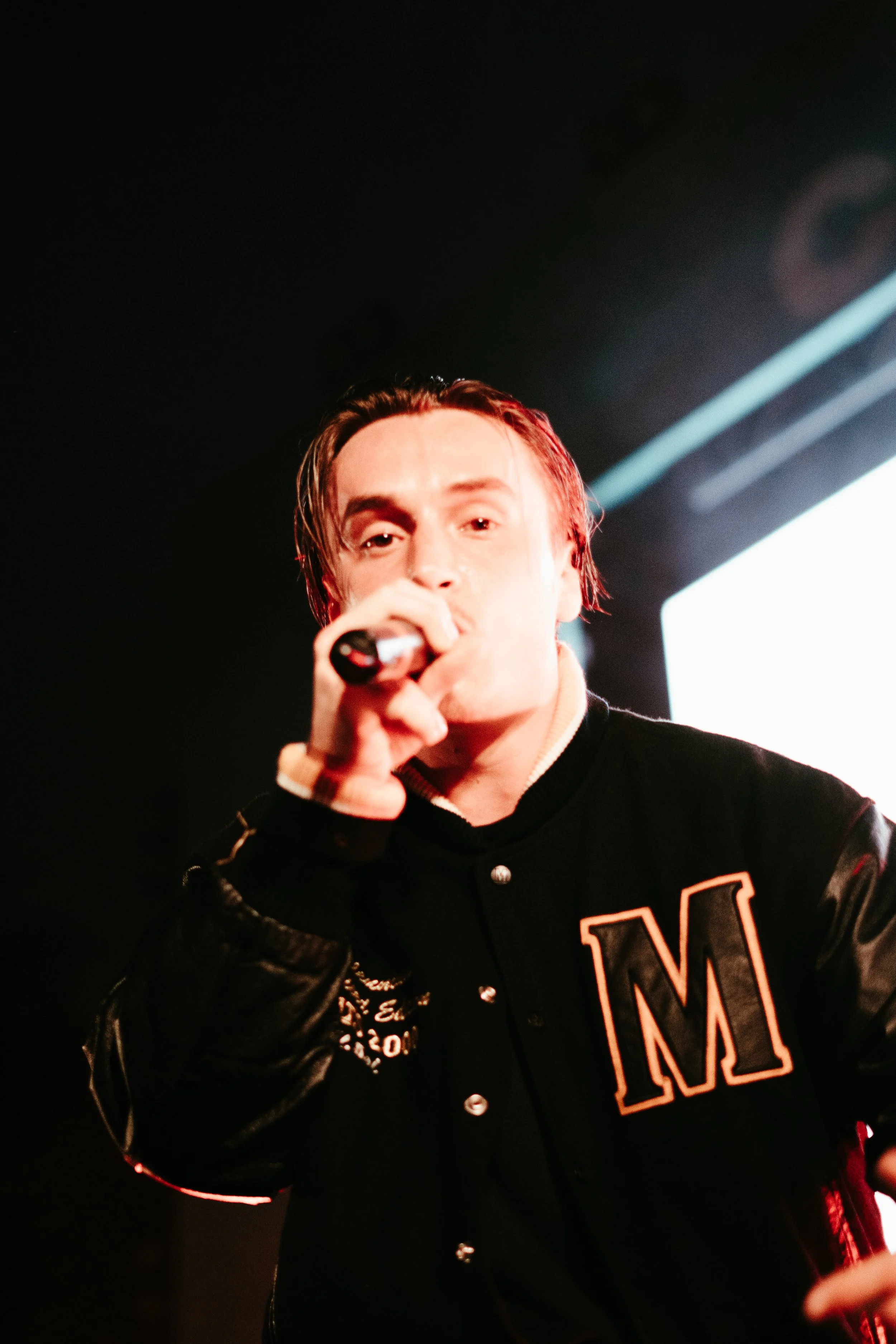 Un homme chante dans un micro lors d'une performance, portant une veste avec la lettre 'M' sur le devant.