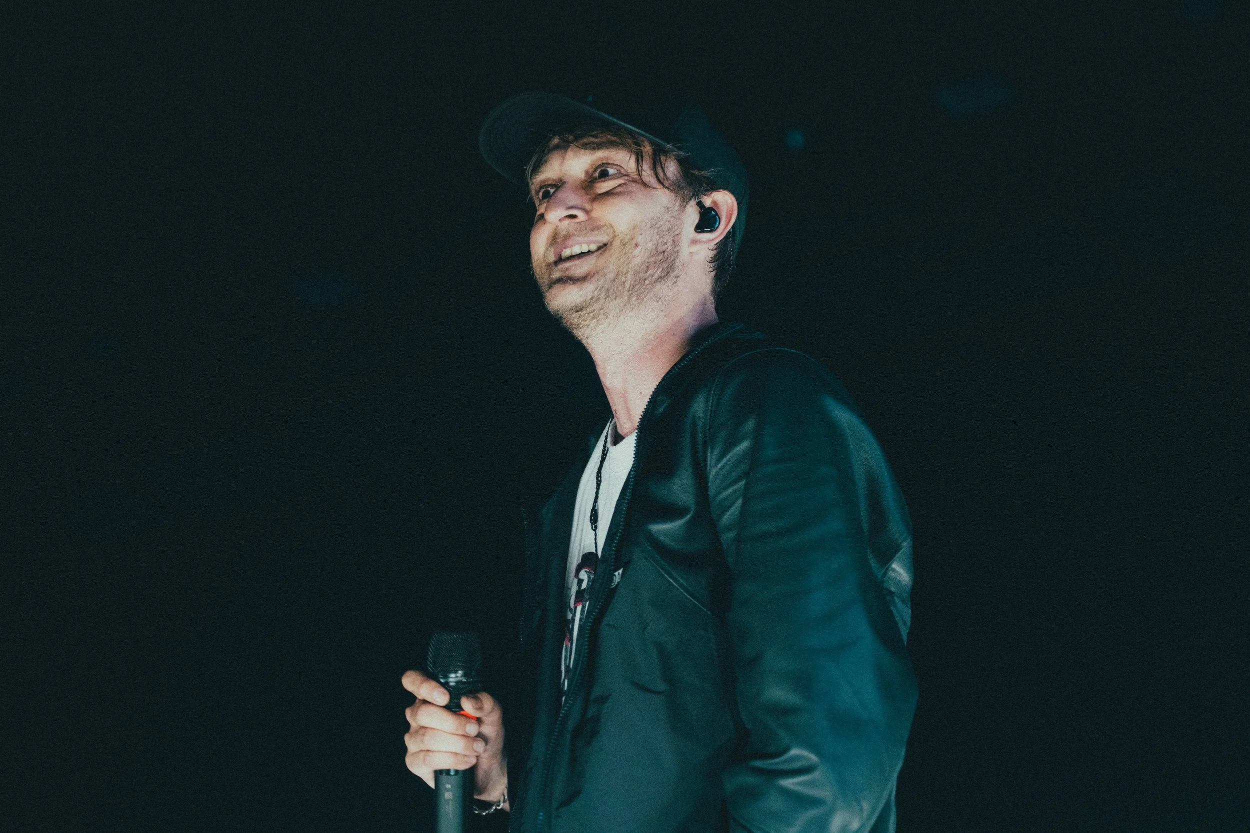 Un homme tenant un micro, portant une casquette et une veste en cuir, souriant et regardant vers la gauche, dans un environnement sombre.