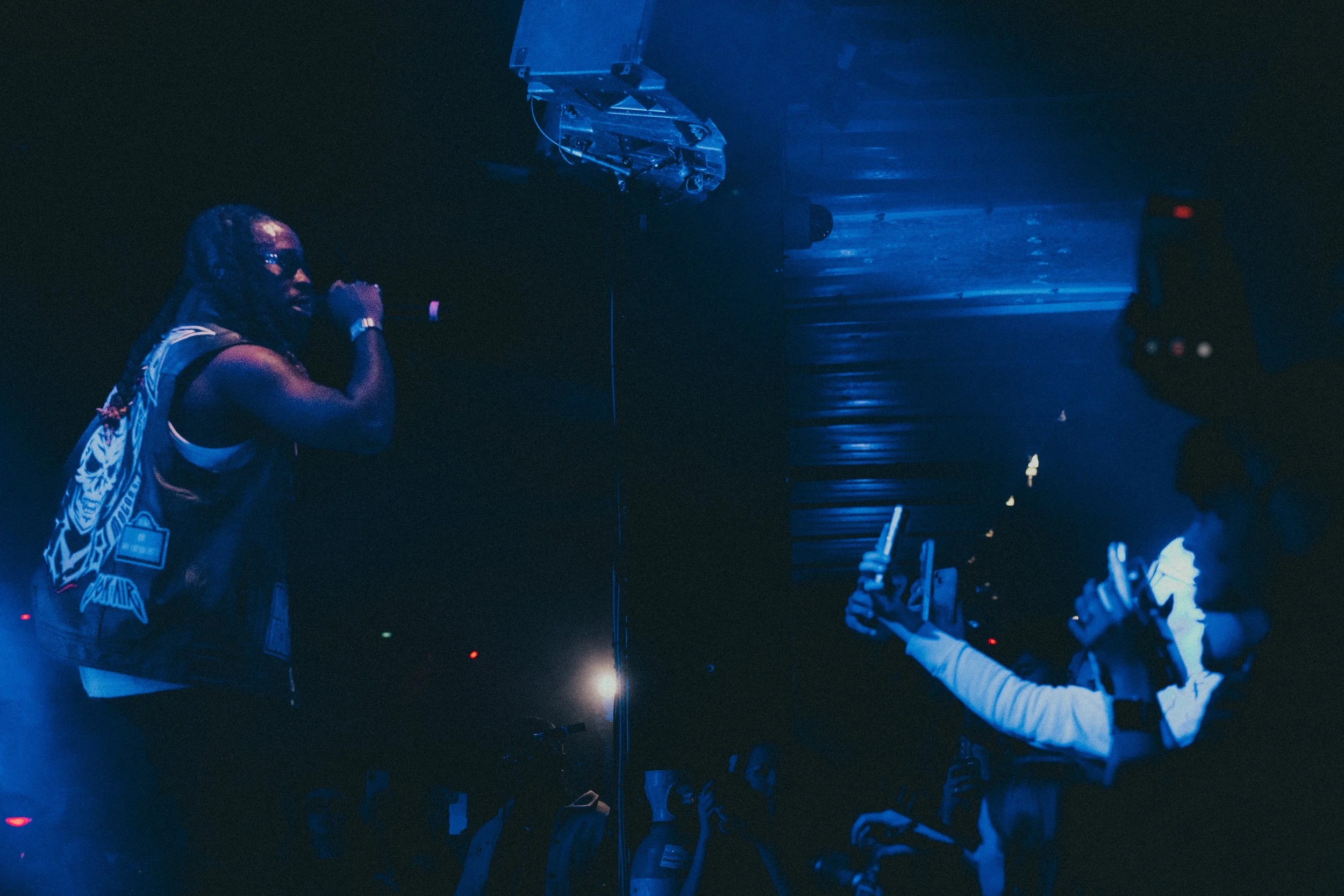 Un homme avec des dreadlocks et un gilet sans manches chante dans un micro sur scène, éclairé par une lumière bleue, avec un public à gauche tenant des téléphones.