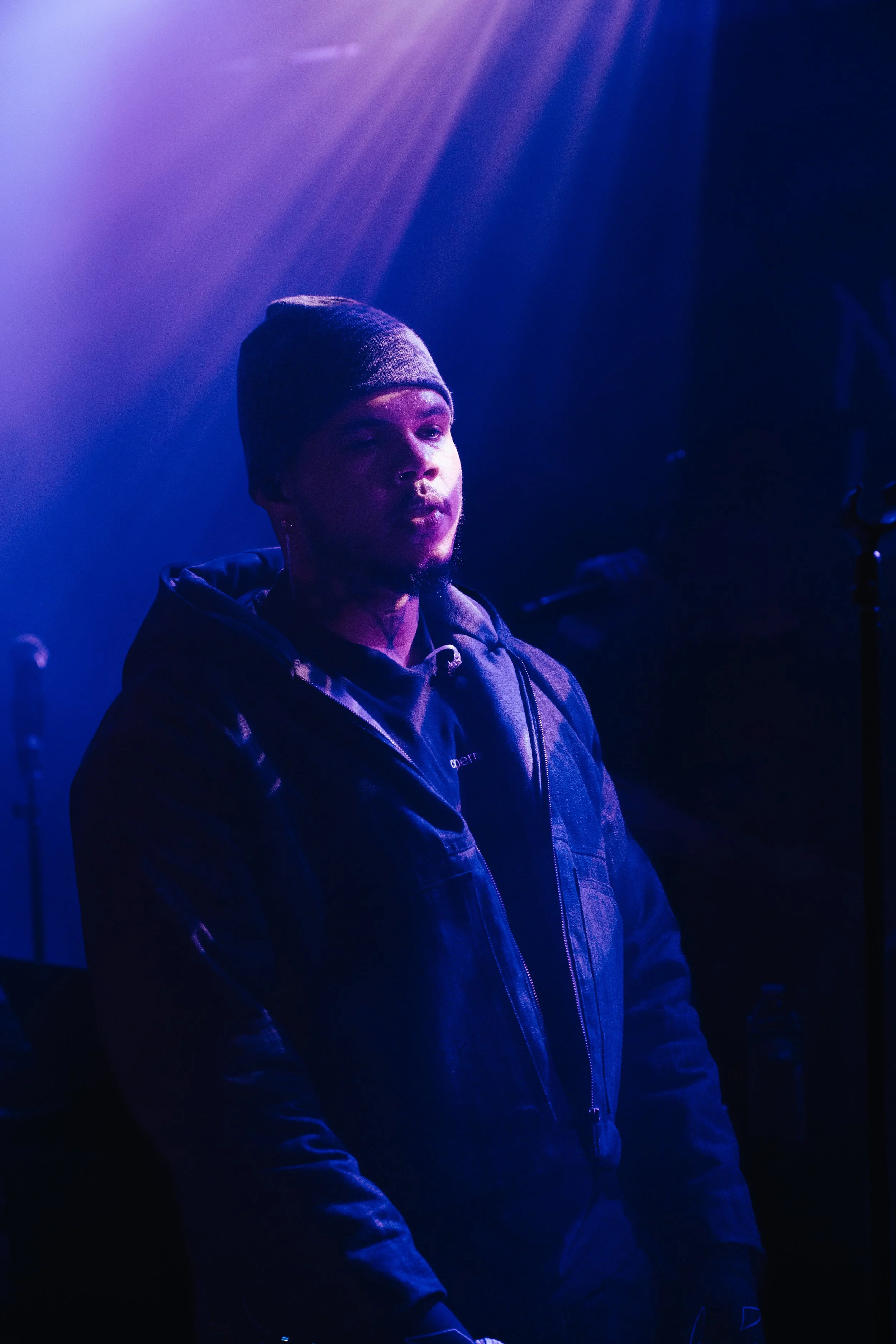 Une personne portant un bonnet gris et une veste noire, dans un environnement sombre éclairé par des lumières violettes et bleues, avec un microphone visible à côté.