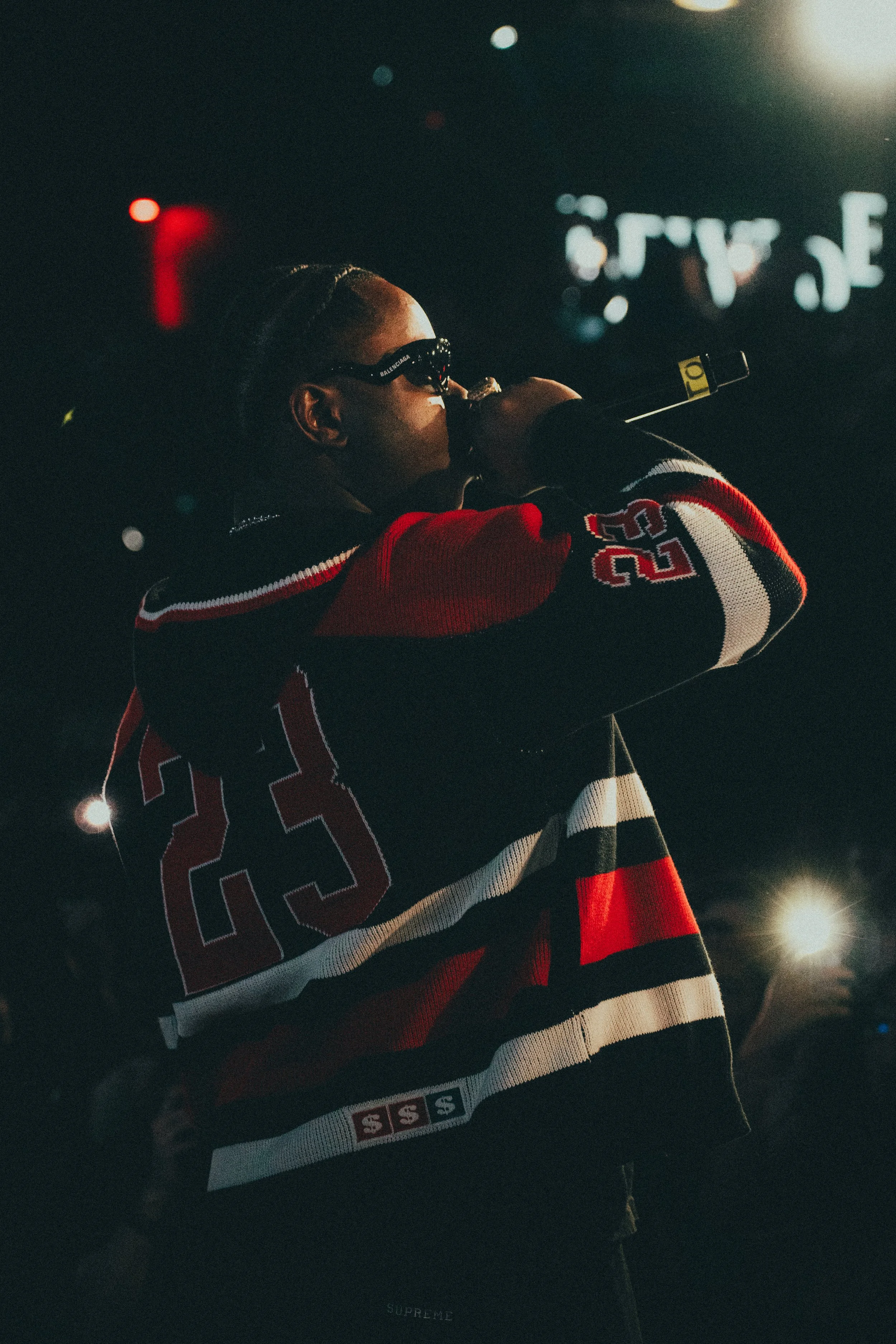 Une personne porte des lunettes de soleil et un sweat à capuche à rayures rouges, noires et blanches, tenant un microphone, dans un environnement sombre avec des lumières floues en arrière-plan.