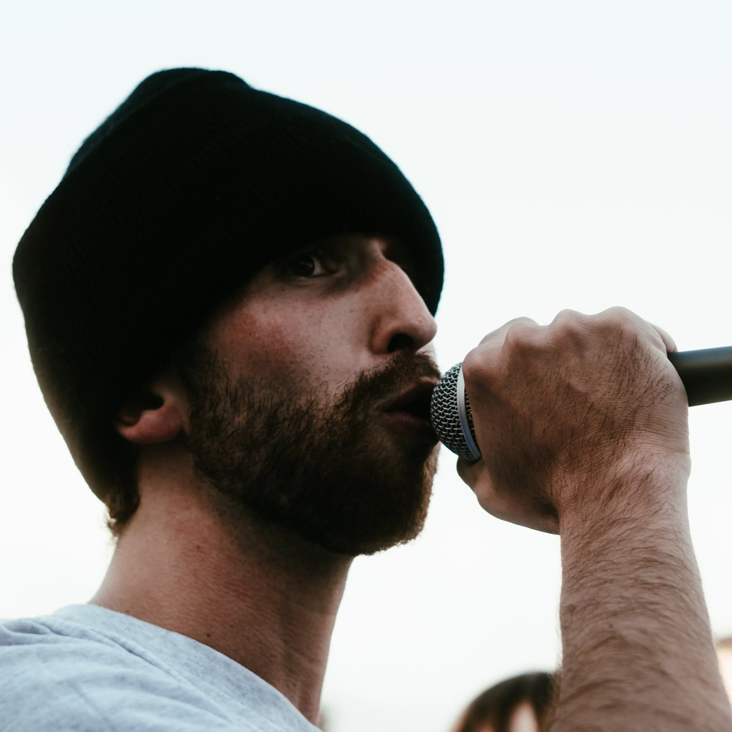 Un homme avec une barbe, portant un bonnet noir, tenant un microphone près de sa bouche, regardant vers l'avant.