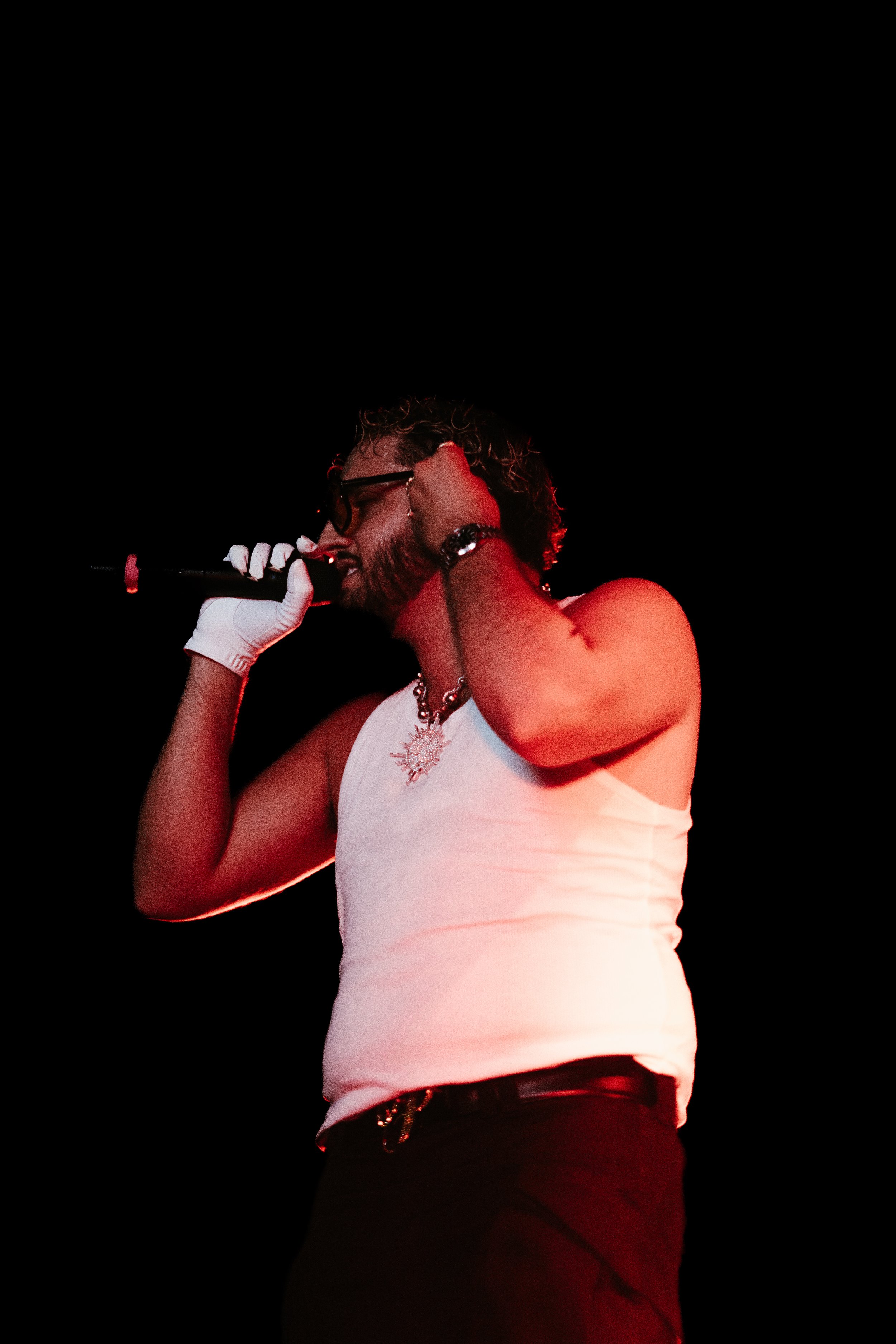 Un homme chante en tenant un micro, portant des lunettes noires, un t-shirt blanc, des bijoux, et une paire de gants blancs sur scène avec un fond noir.
