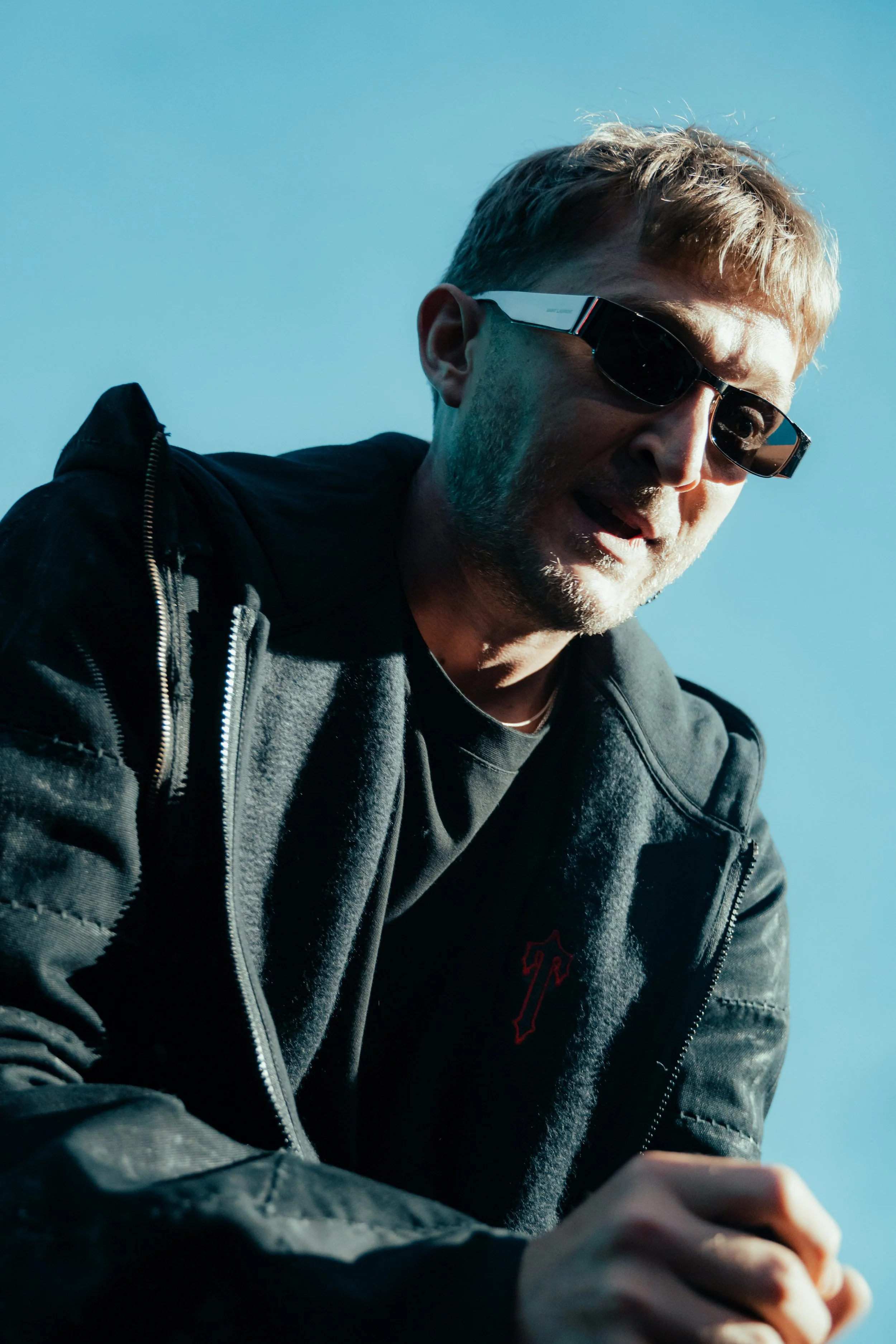 Un homme portant des lunettes de soleil noires et une veste noire, regardant vers la caméra contre un ciel bleu.