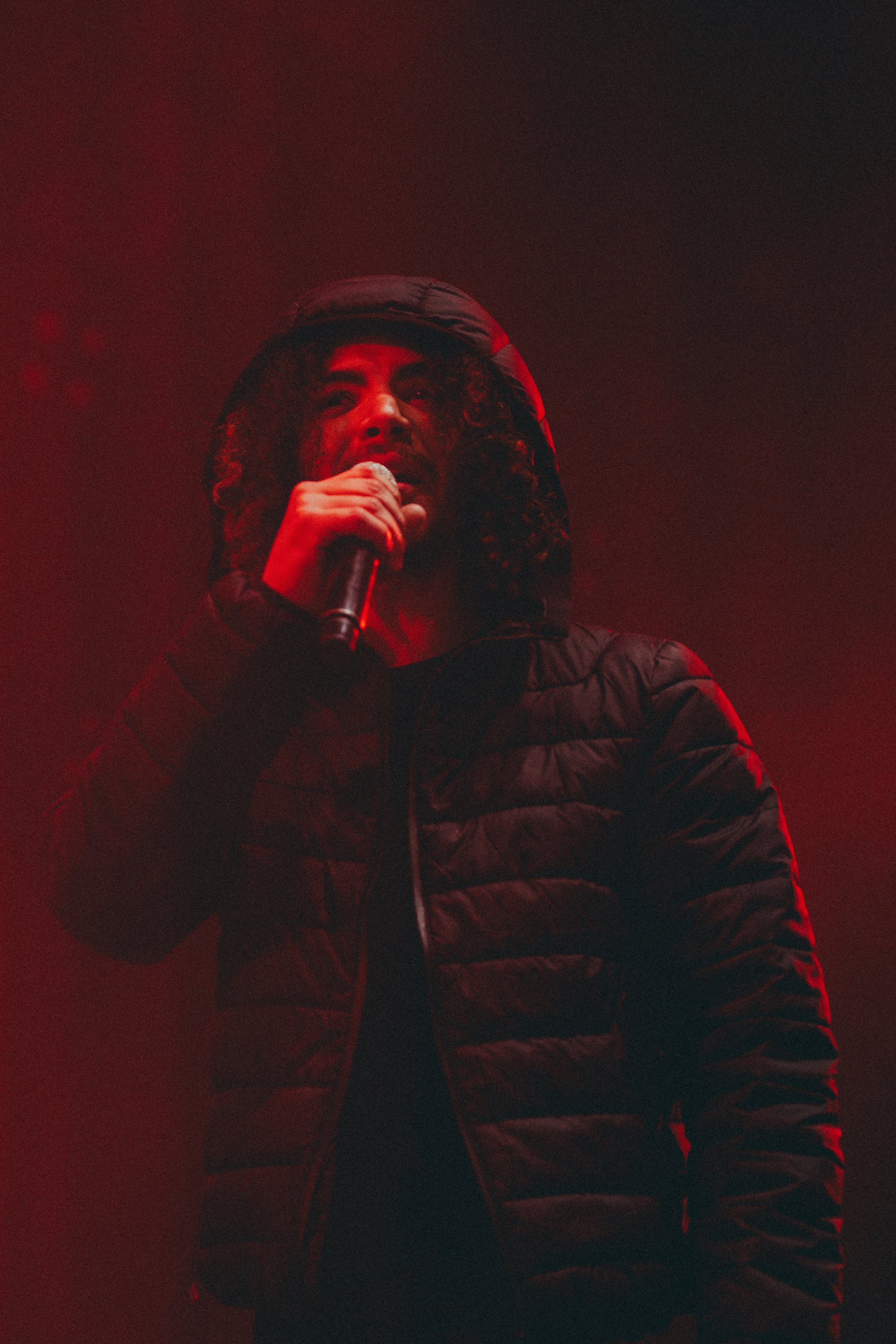 Un homme portant une veste noire tient un microphone dans la main, éclairé par une lumière rouge, sur scène.