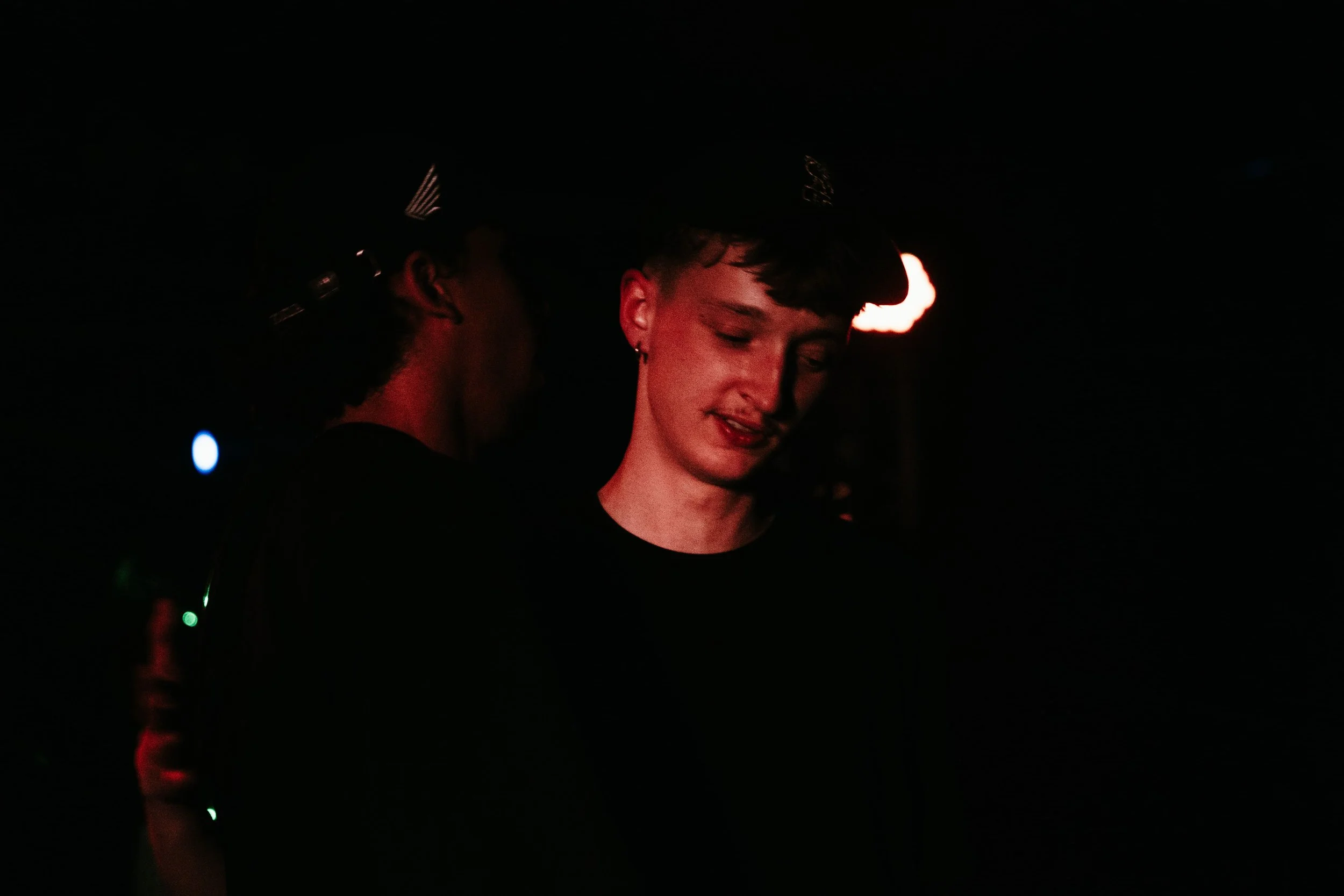 Deux personnes dans une pièce sombre, l'une regarde vers le bas, l'autre regarde légèrement de côté, tous deux portants des casquettes. La scène est faiblement éclairée avec des lumières rouges et bleues en arrière-plan.