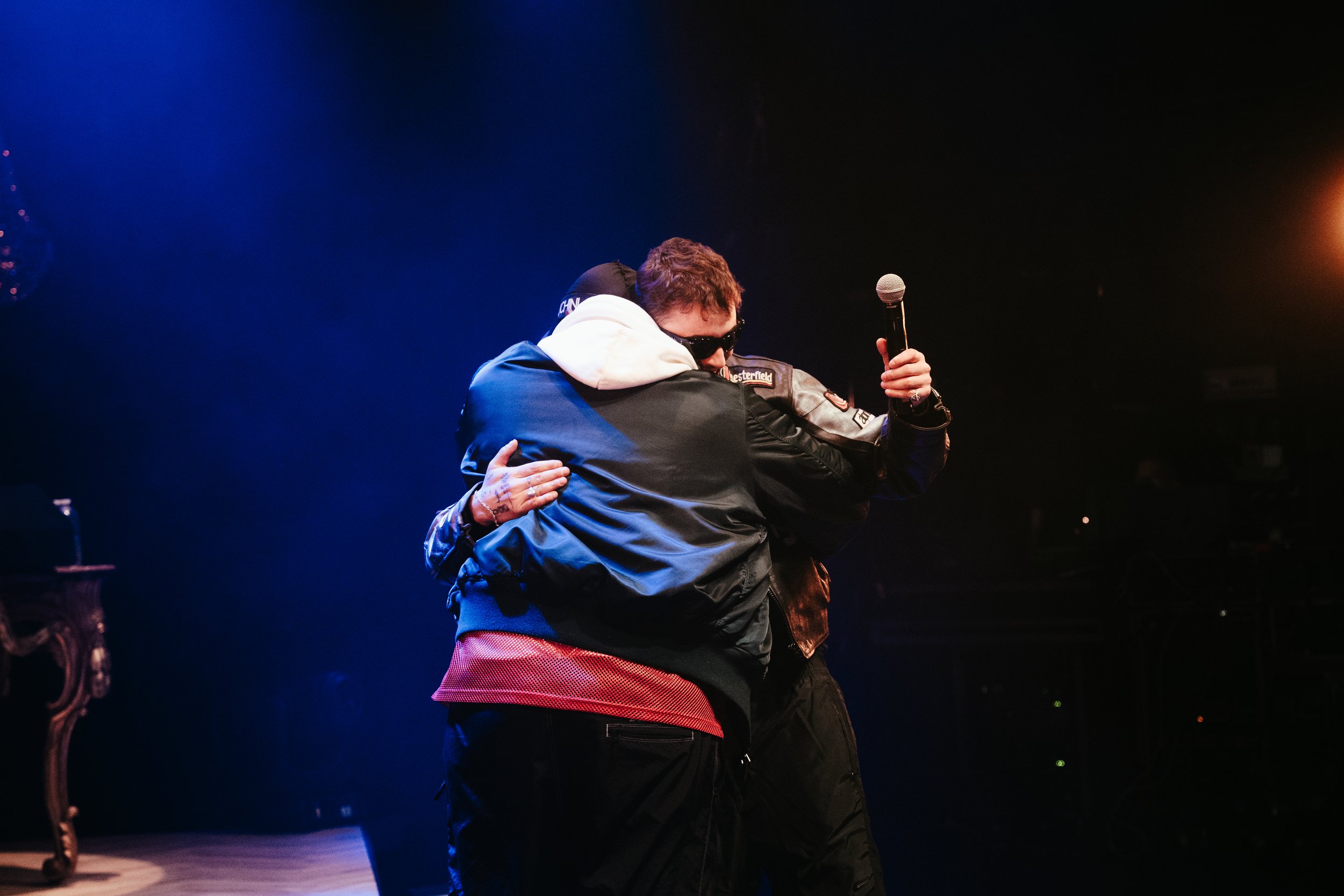 Deux personnes s'embrassent sur scène, une tient un micro, dans une ambiance sombre et éclairée par des lumières chaudes.