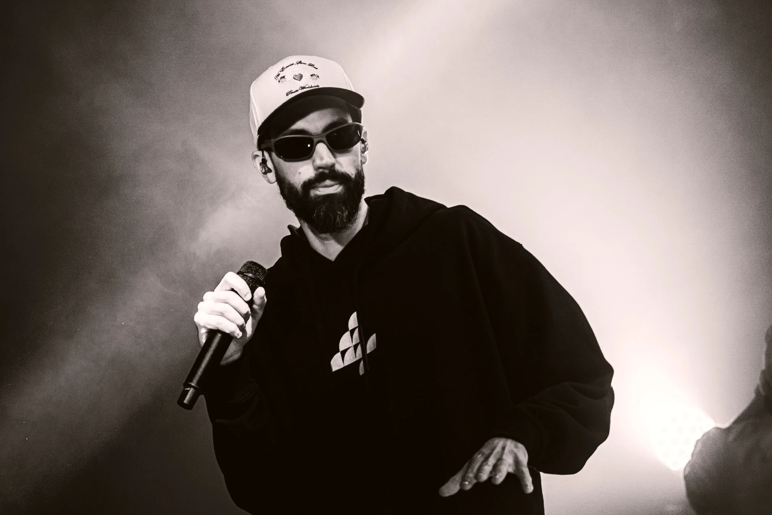Un homme avec une barbe, portant des lunettes de soleil, un chapeau blanc, une veste noire et un sweat noir, tient un micro sur scène dans un contexte sombre.