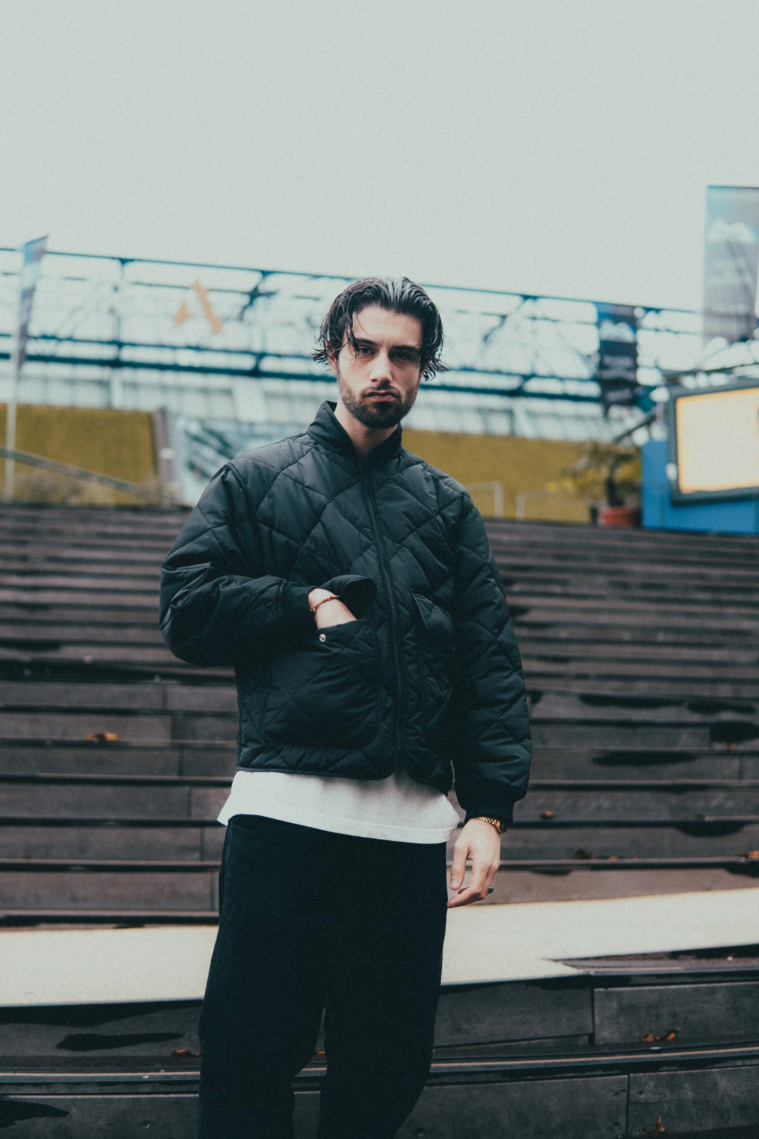 Jeune homme avec des cheveux noirs ondulés, portant une veste noire en quilté, debout devant des gradins en bois dans un environnement urbain.