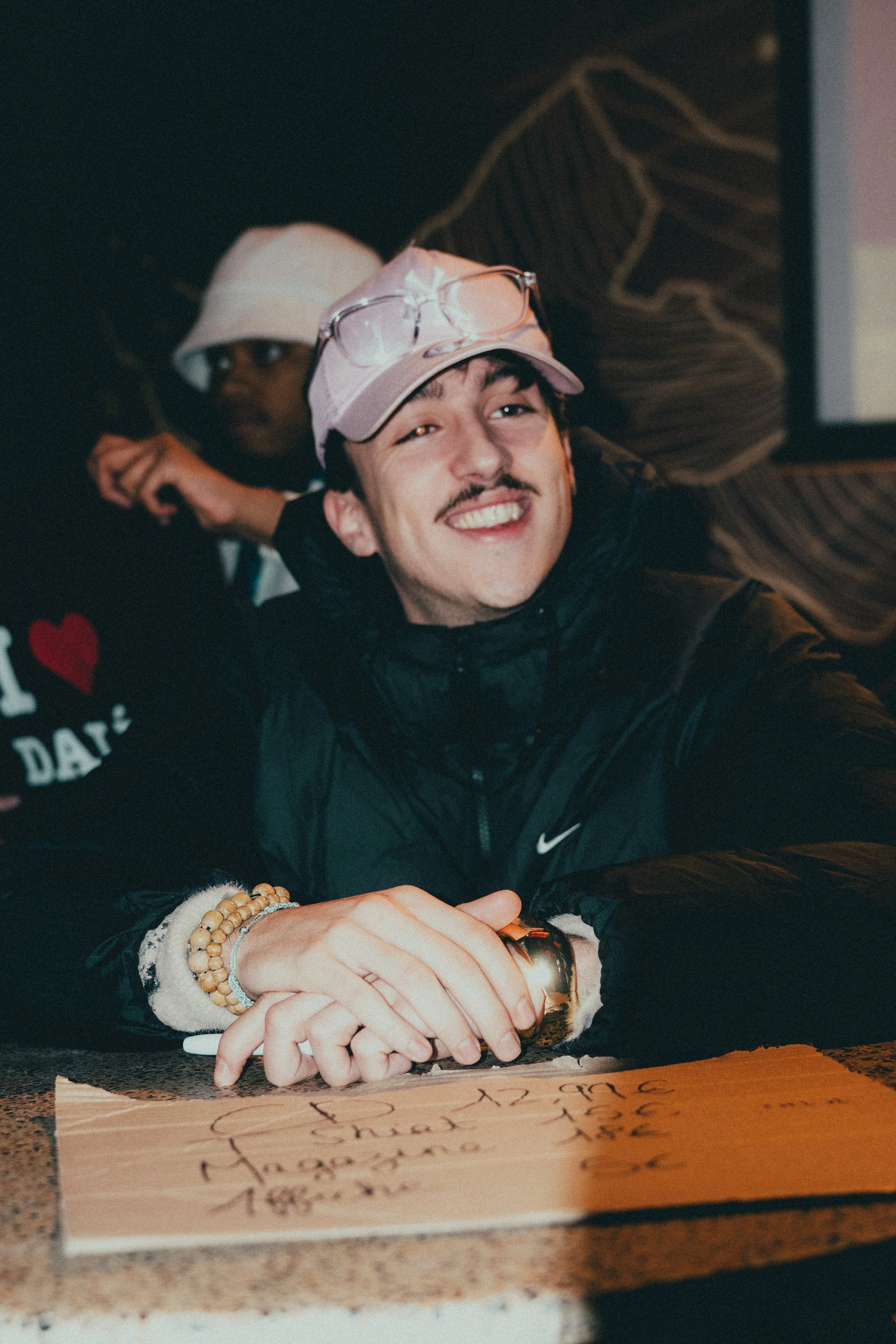 Un homme souriant, portant une casquette rose, des lunettes sur la tête, assis à une table avec une carte ou un morceau de papier devant lui, dans un environnement intérieur