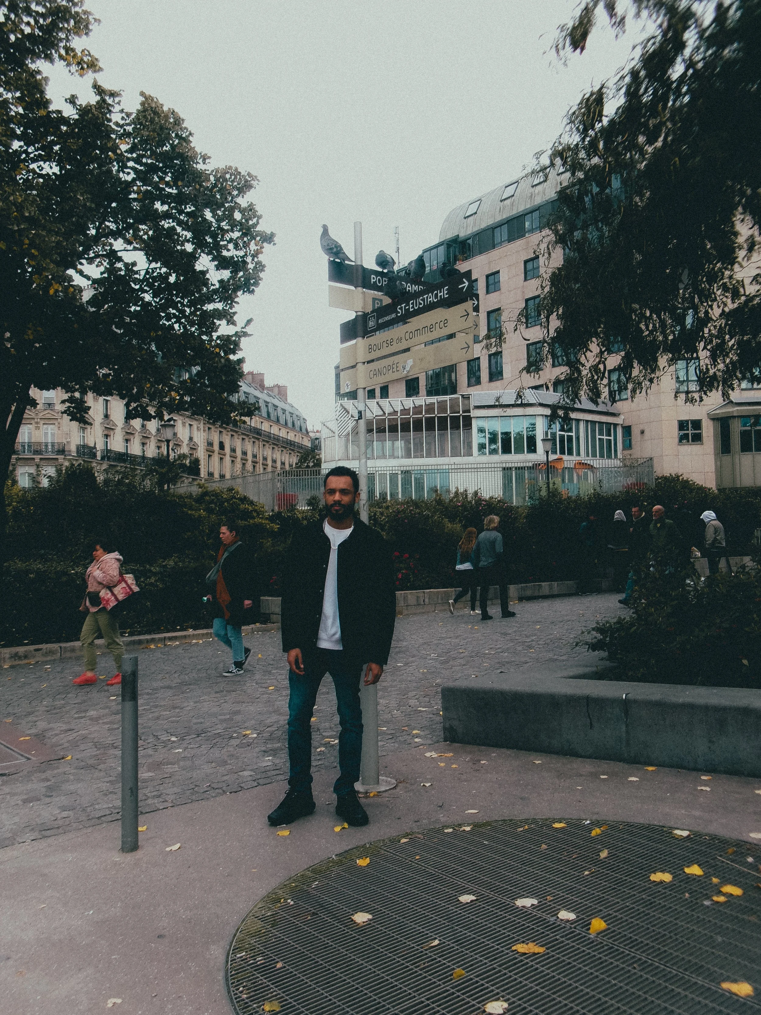 Une scène urbaine avec plusieurs personnes dans un parc, un homme debout en avant-plan, des bâtiments modernes en arrière-plan, un panneau de signalisation avec plusieurs flèches, des arbres et des feuilles tombées au sol.