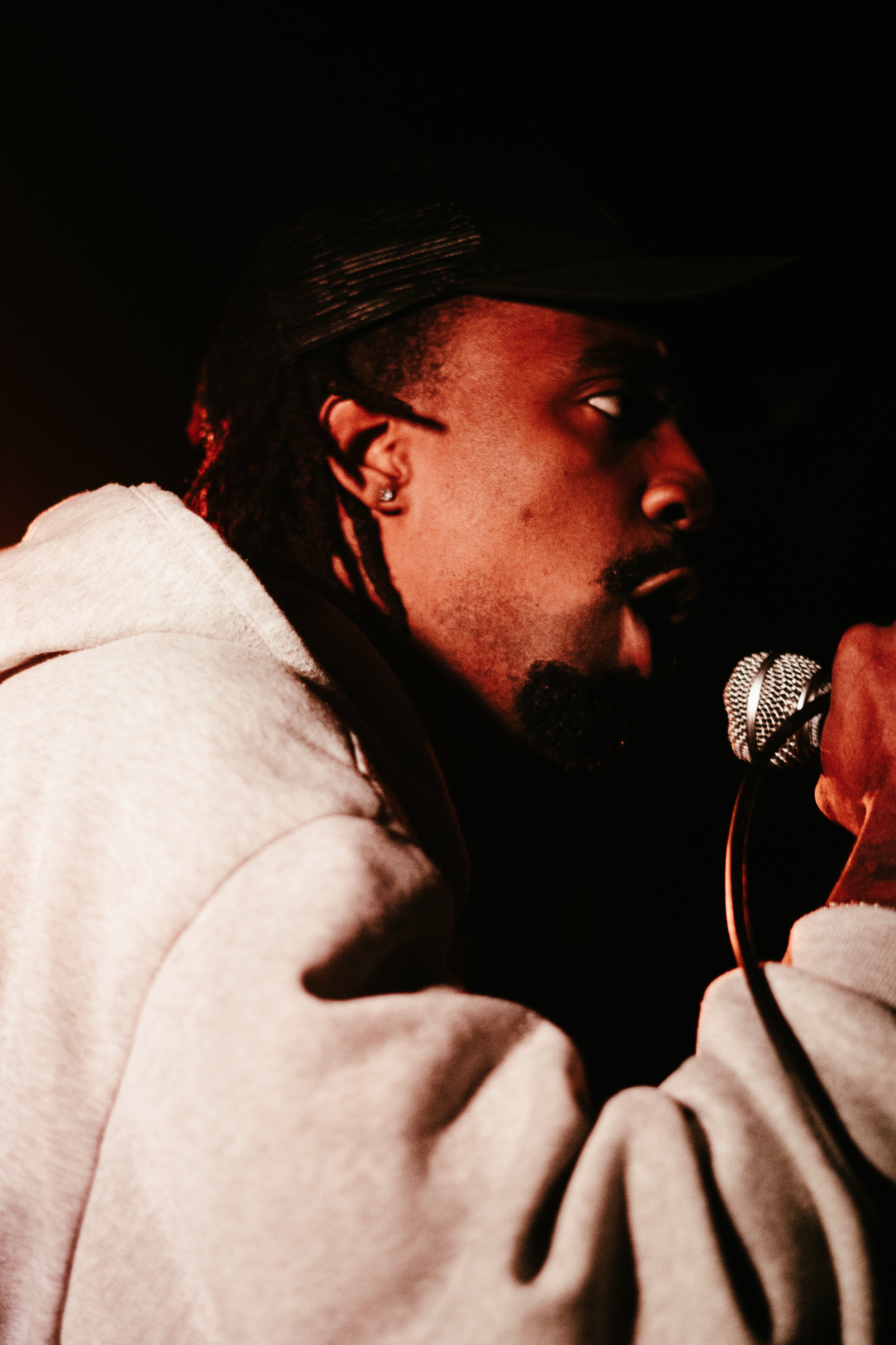 Un homme avec des cheveux en dreadlocks, portant une casquette, tenant un microphone et semble en train de chanter ou de parler dans un environnement sombre.