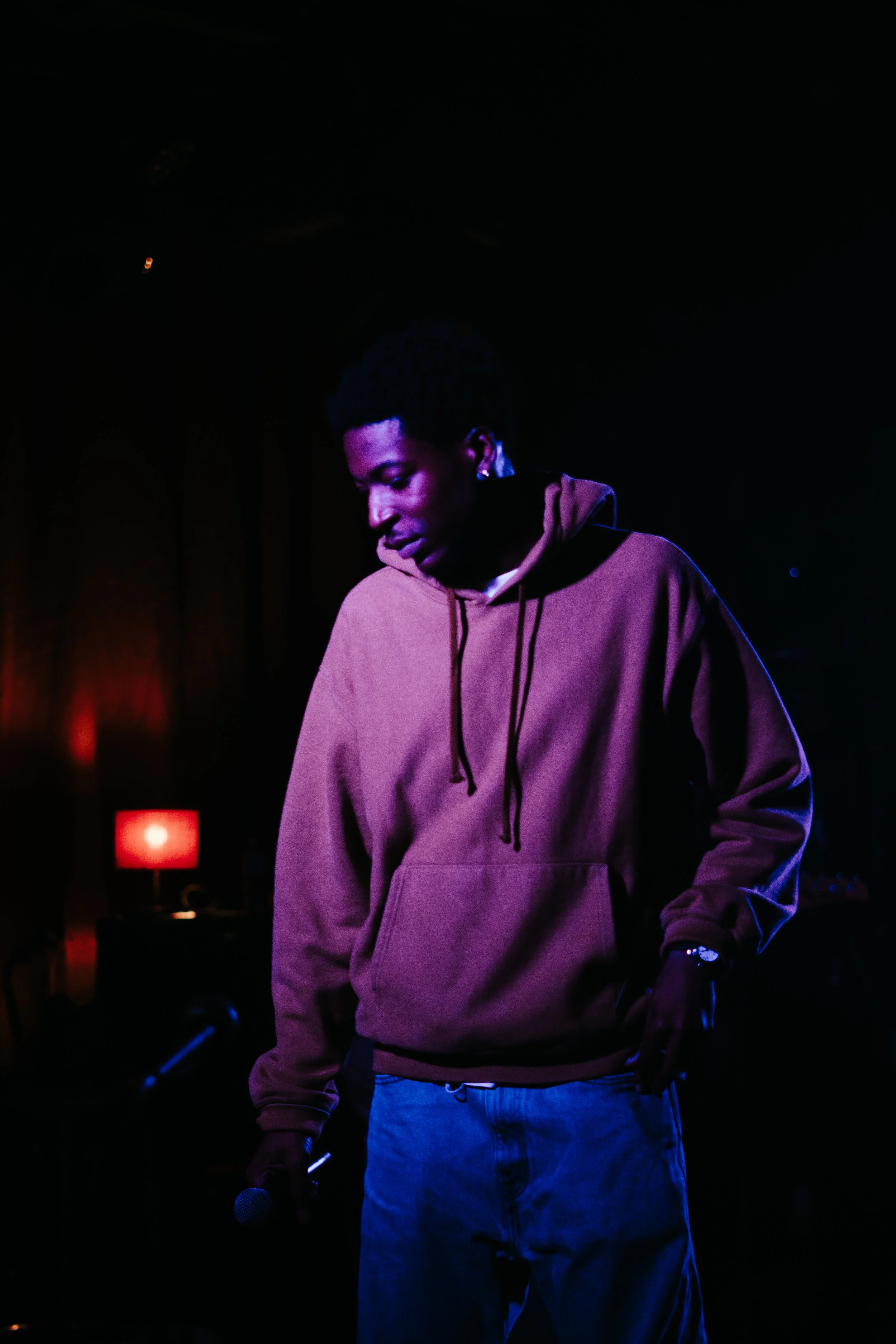 Un jeune homme tenant un micro, portant un sweat à capuche marron, dans un environnement sombre avec une lumière rouge en fond.