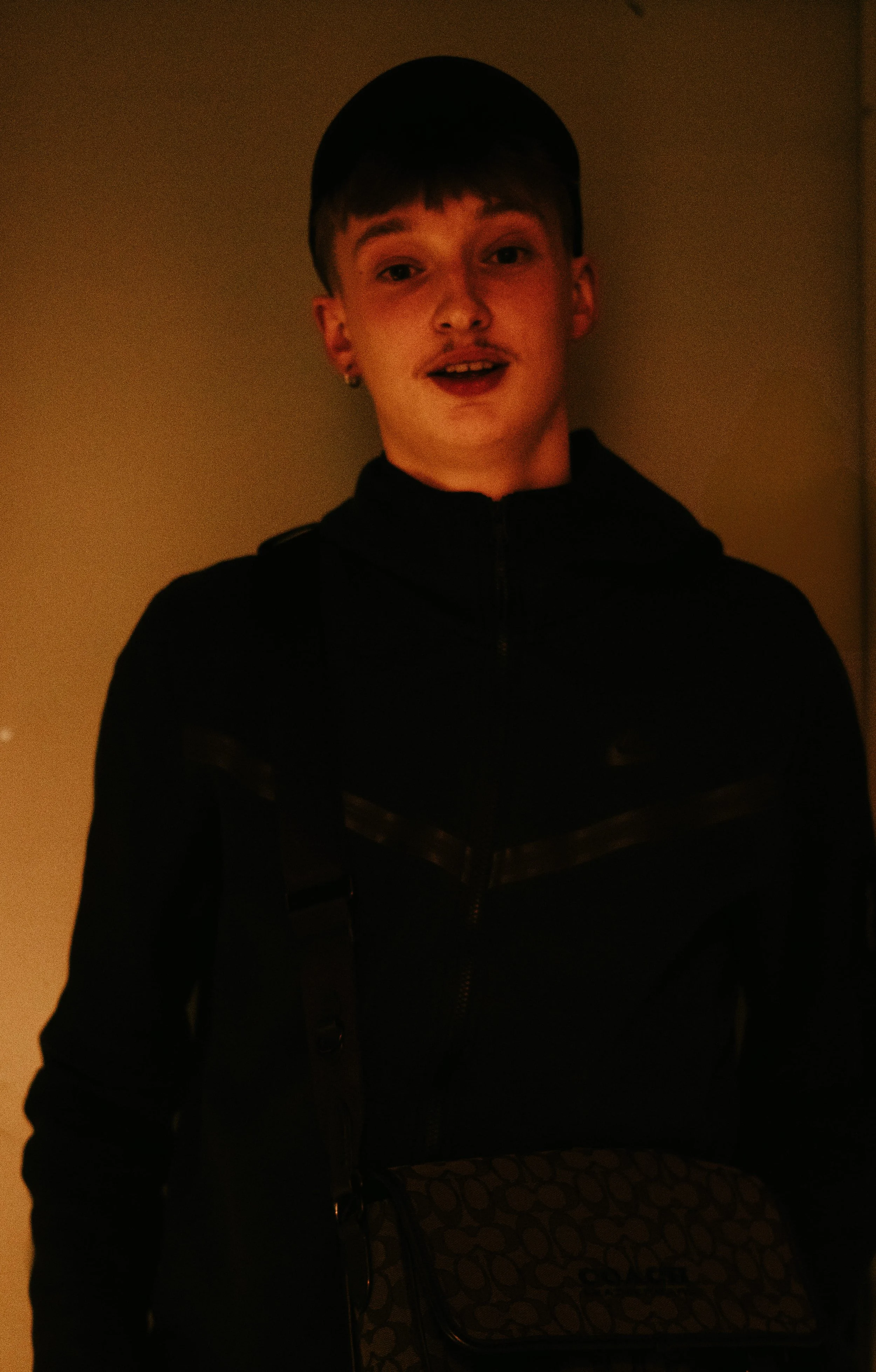 Jeune homme souriant portant une casquette noire, en tenue noire avec un sac à main à motifs, dans une ambiance sombre.