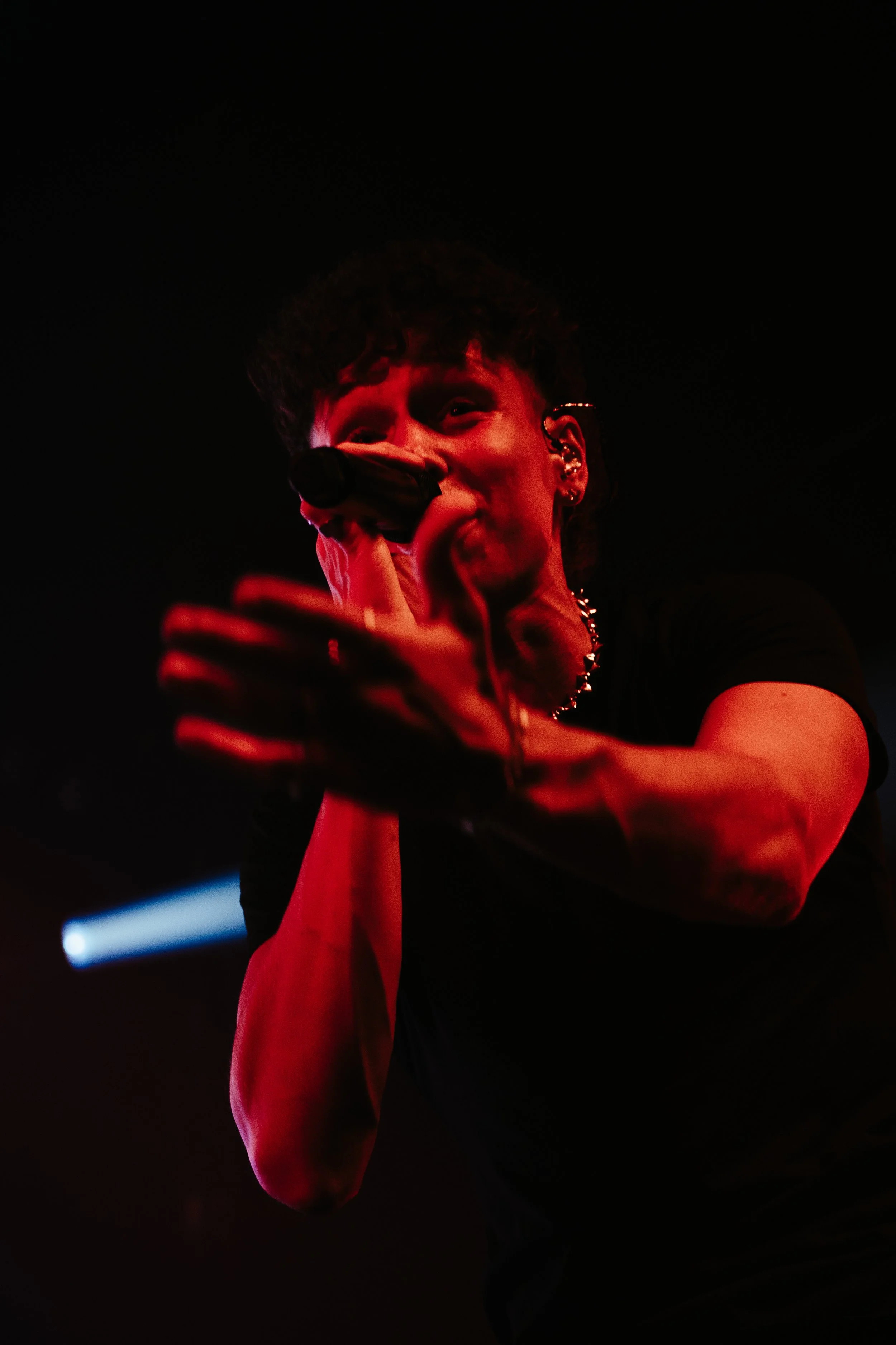 Un chanteur masculine avec des piercings, tenant un microphone, en train de chanter lors d'un concert, éclairé par un éclairage rouge et noir avec un faisceau de lumière en fond.