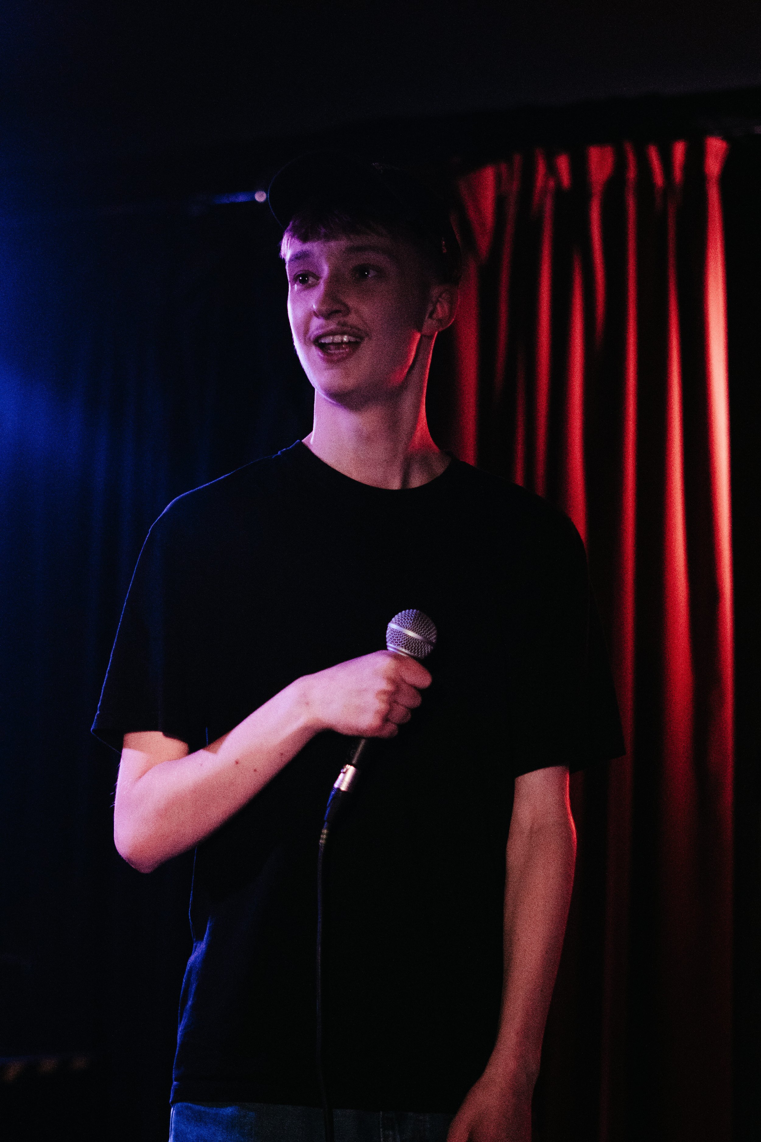 Jeune homme tenant un microphone sur scène, avec des rideaux rouges en arrière-plan.