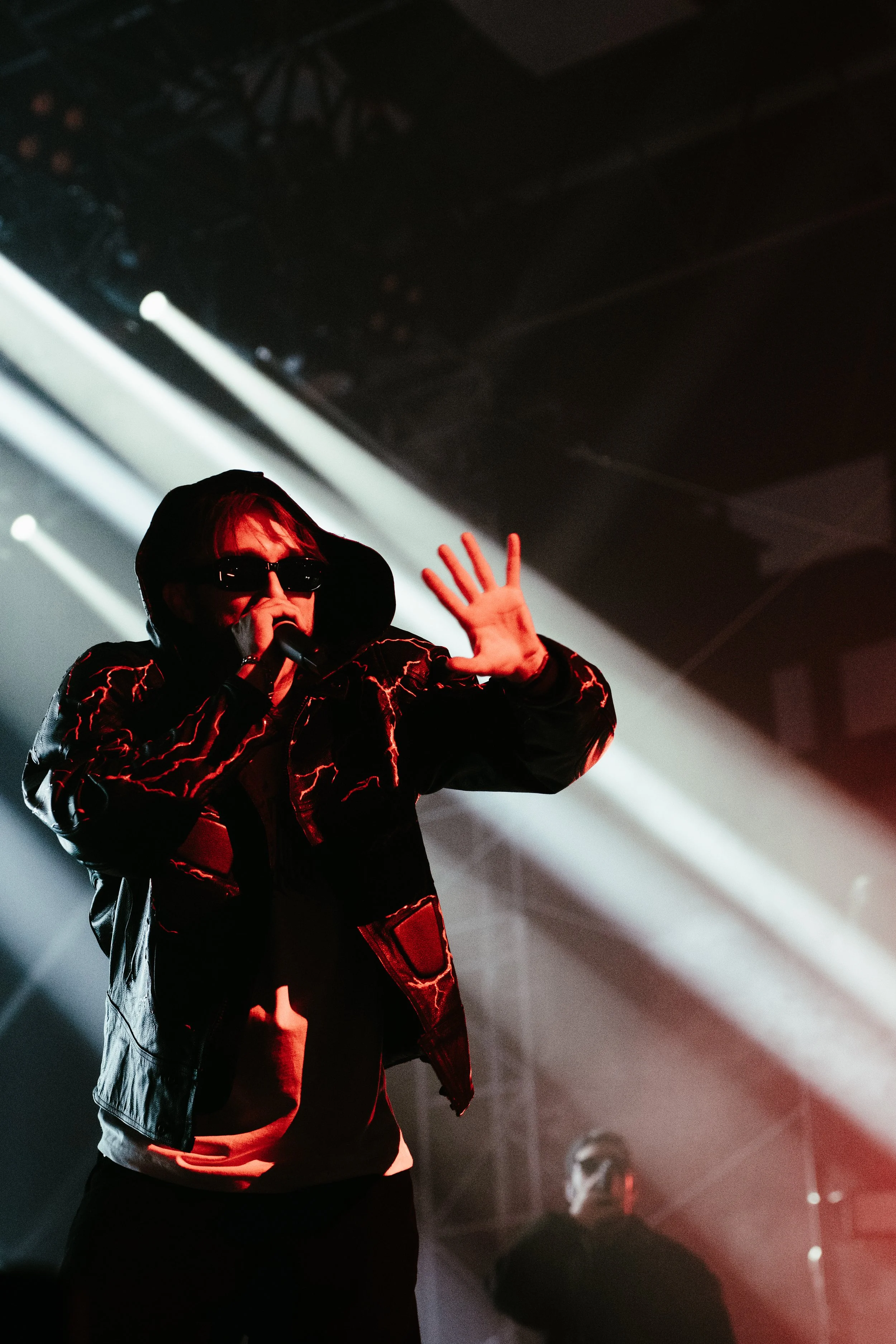 Un artiste en tenue sombre, portant des lunettes de soleil et un sac à main, chante en microphone sur scène avec des lumières de concert en arrière-plan.