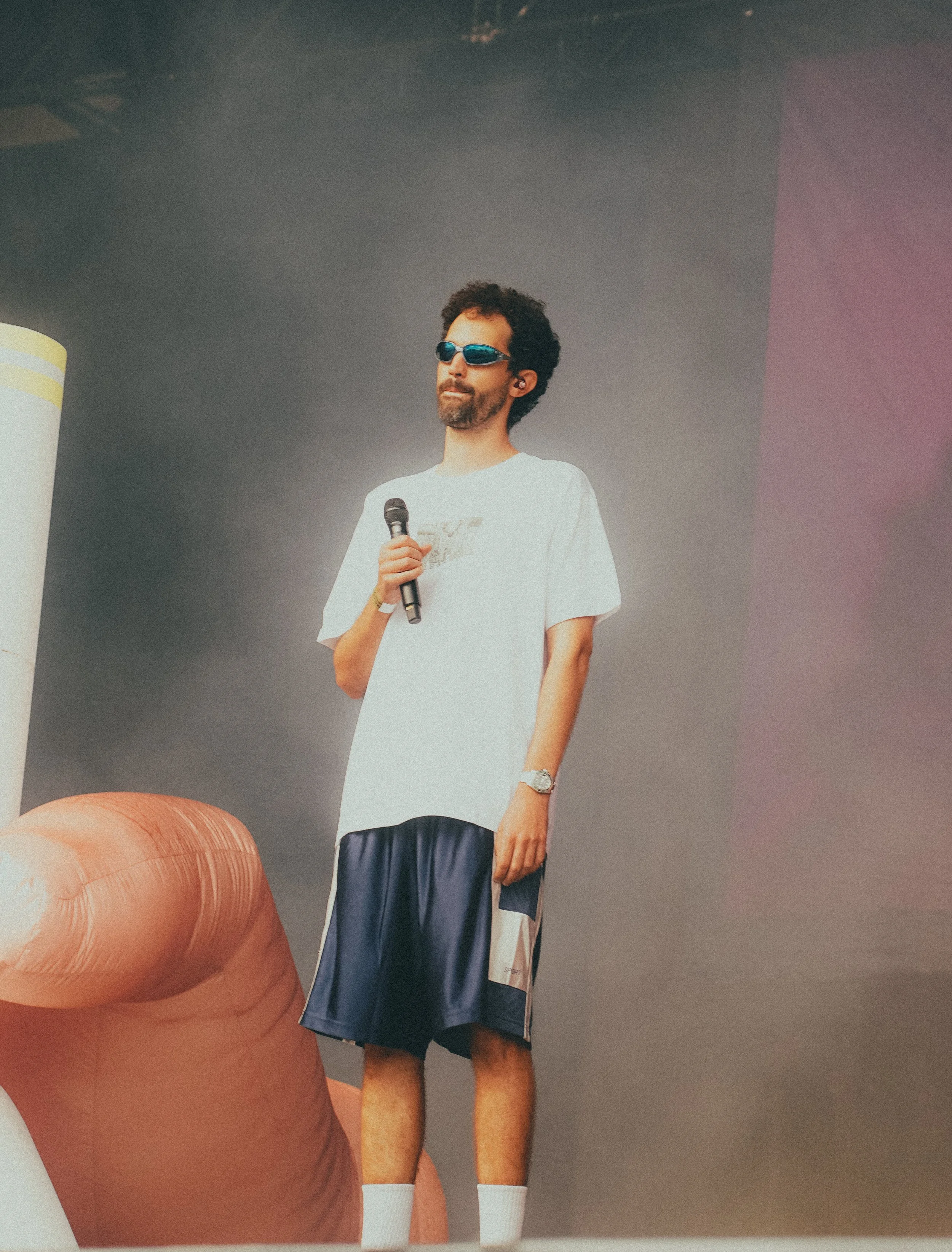 Un homme debout en tenue décontractée, portant des lunettes de soleil bleues, tenant un microphone, dans un environnement intérieur avec un fond gris et un canapé en cuir marron à proximité.
