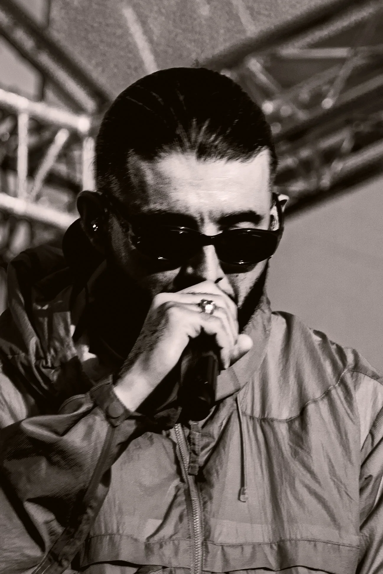 Homme portant des lunettes sombres, un sweat à capuche et un blouson, tenant un microphone et semblant chanter ou parler.