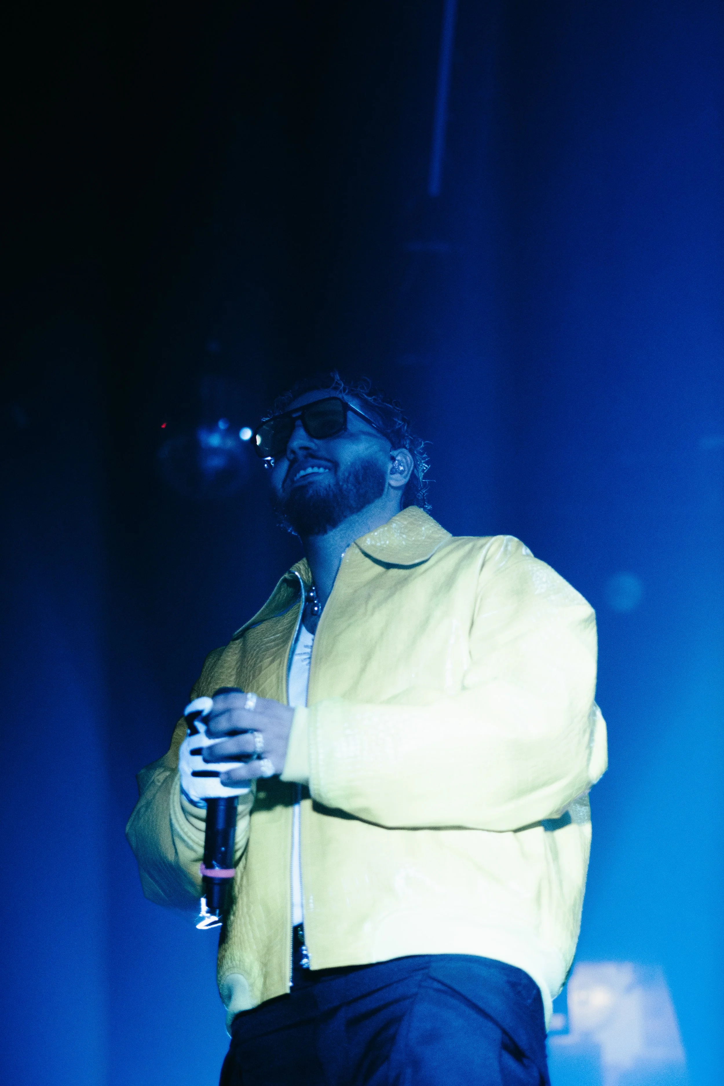 Un homme souriant en portant des lunettes de soleil et un blouson jaune, tenant un micro sur scène avec un éclairage bleu.