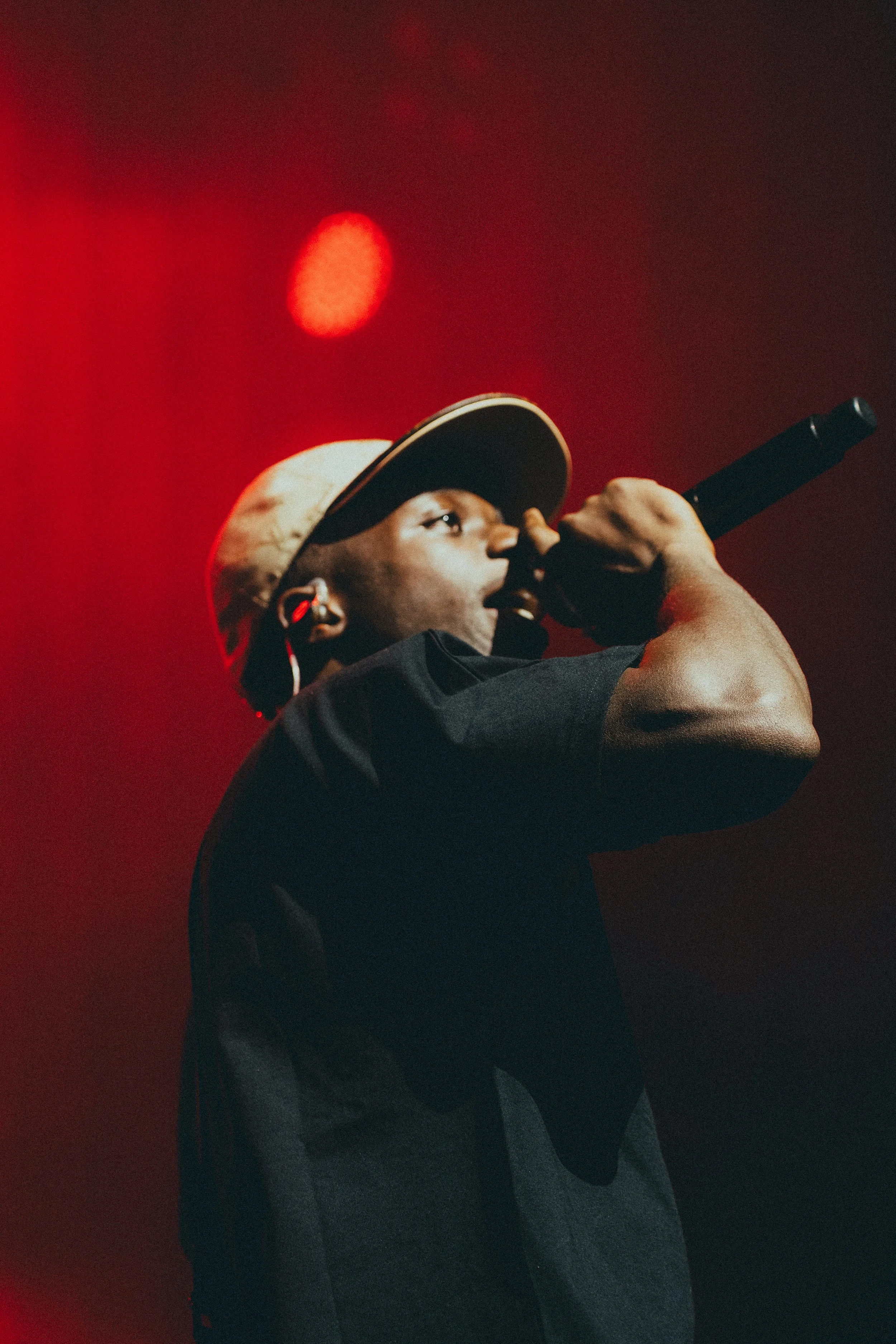 Un homme qui chante ou rappe dans un microphone, portant une casquette beige, avec un fond rouge.