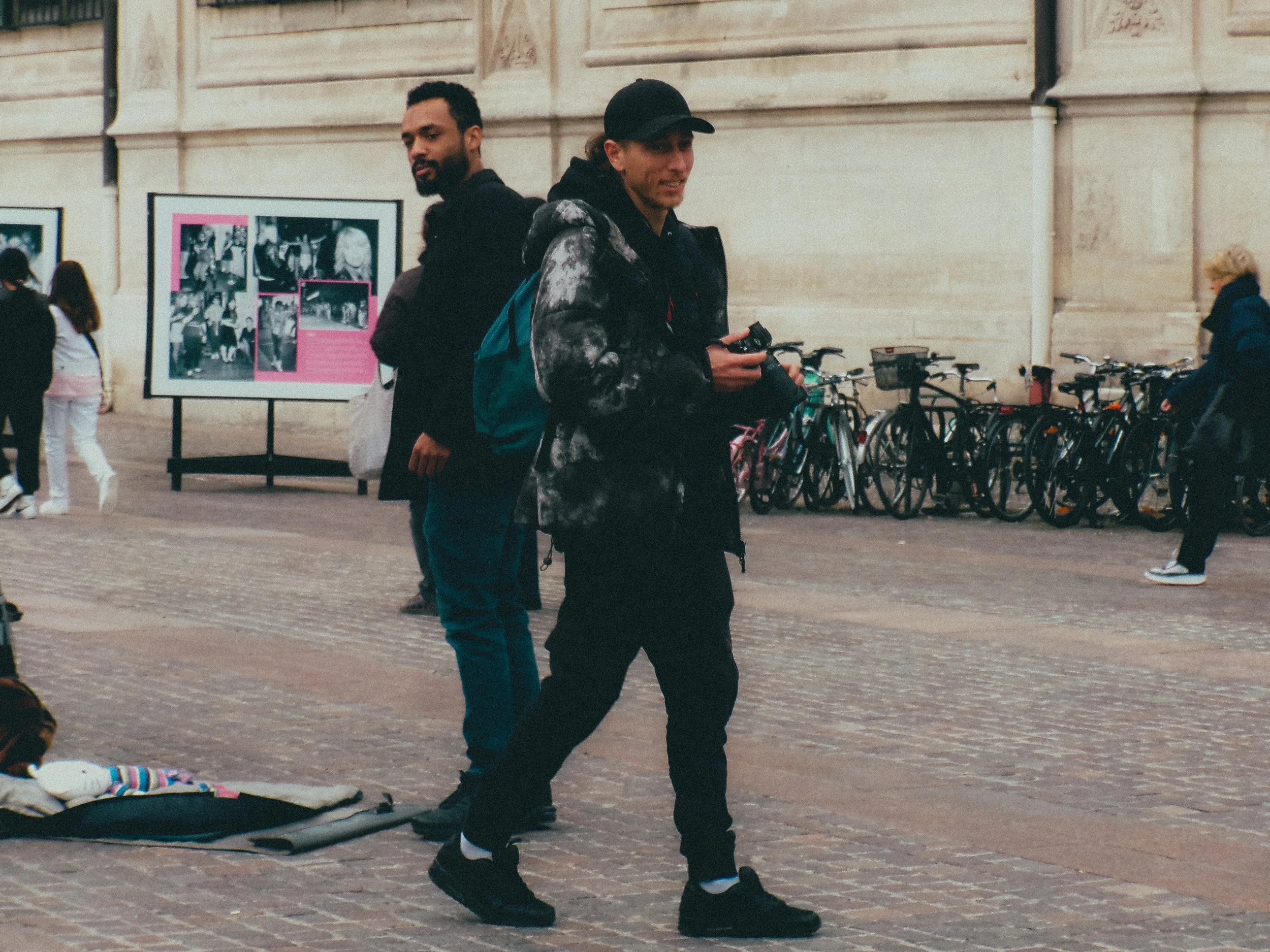 Deux jeunes hommes se tiennent dans la rue près de vélos stationnés contre un mur, un d'eux tient un appareil photo, dans une zone urbaine avec des passants et un panneau d'affichage en arrière-plan.