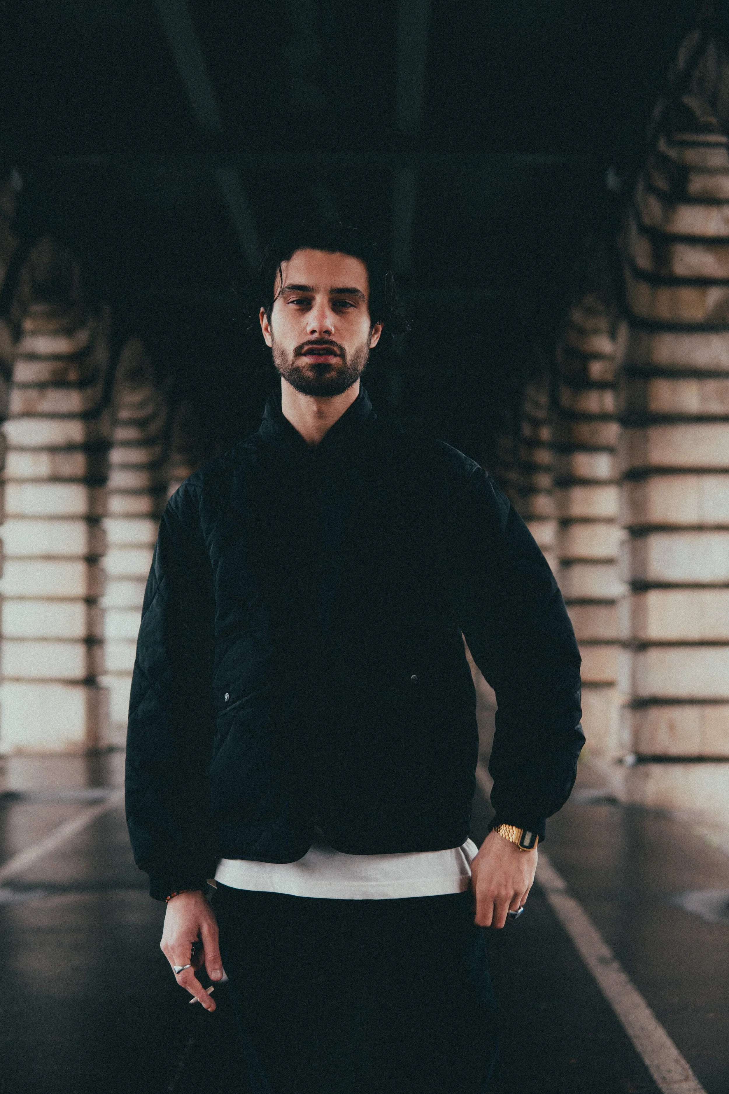 Jeune homme avec une barbe, portant une veste noire, dans un parking sous un pont avec des piles de briques ou de pierre en arrière-plan.