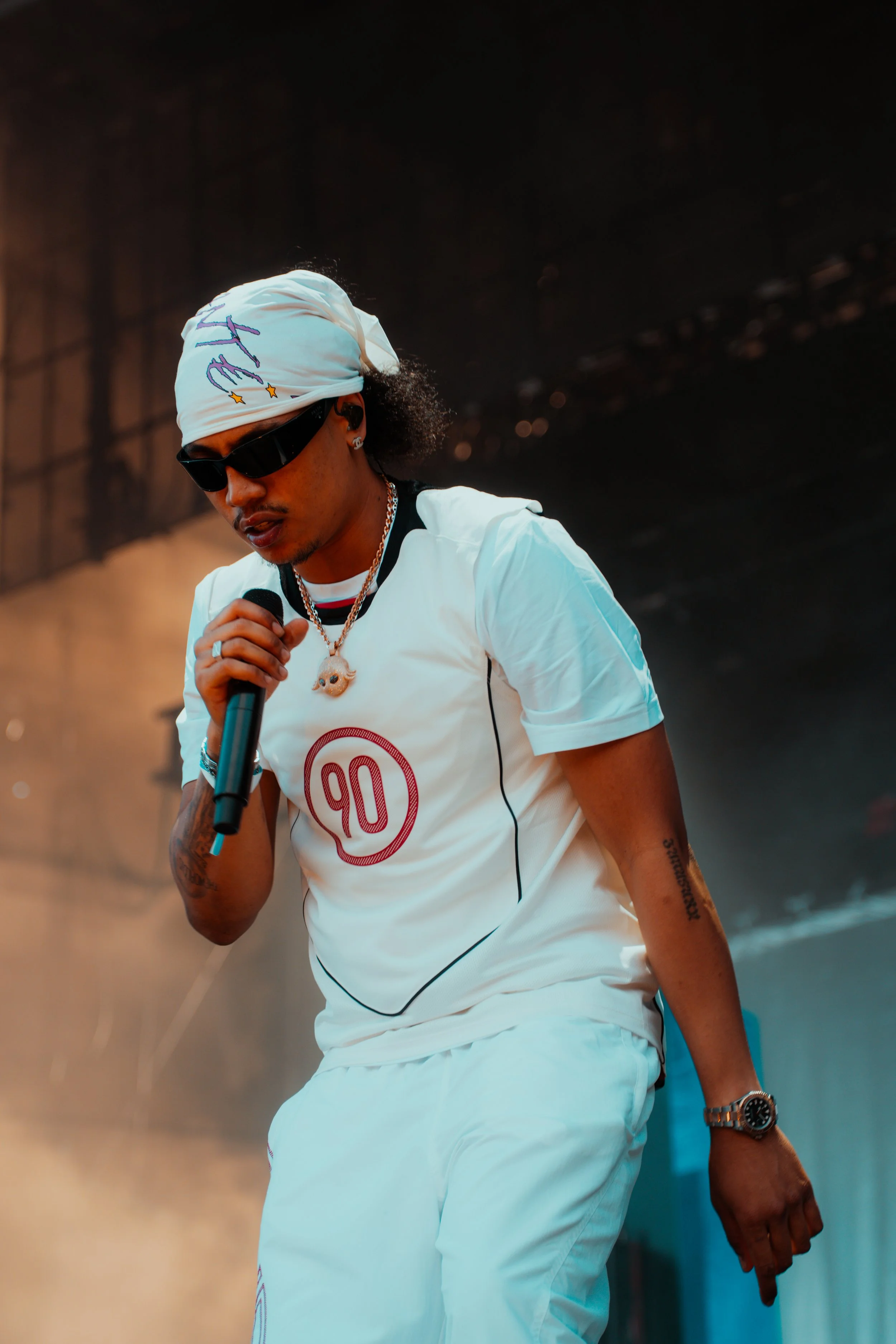 Un homme portant des vêtements blancs, un bandana blanc, des lunettes de soleil noires, tenant un microphone, avec des tatouages, des bijoux et une montre.