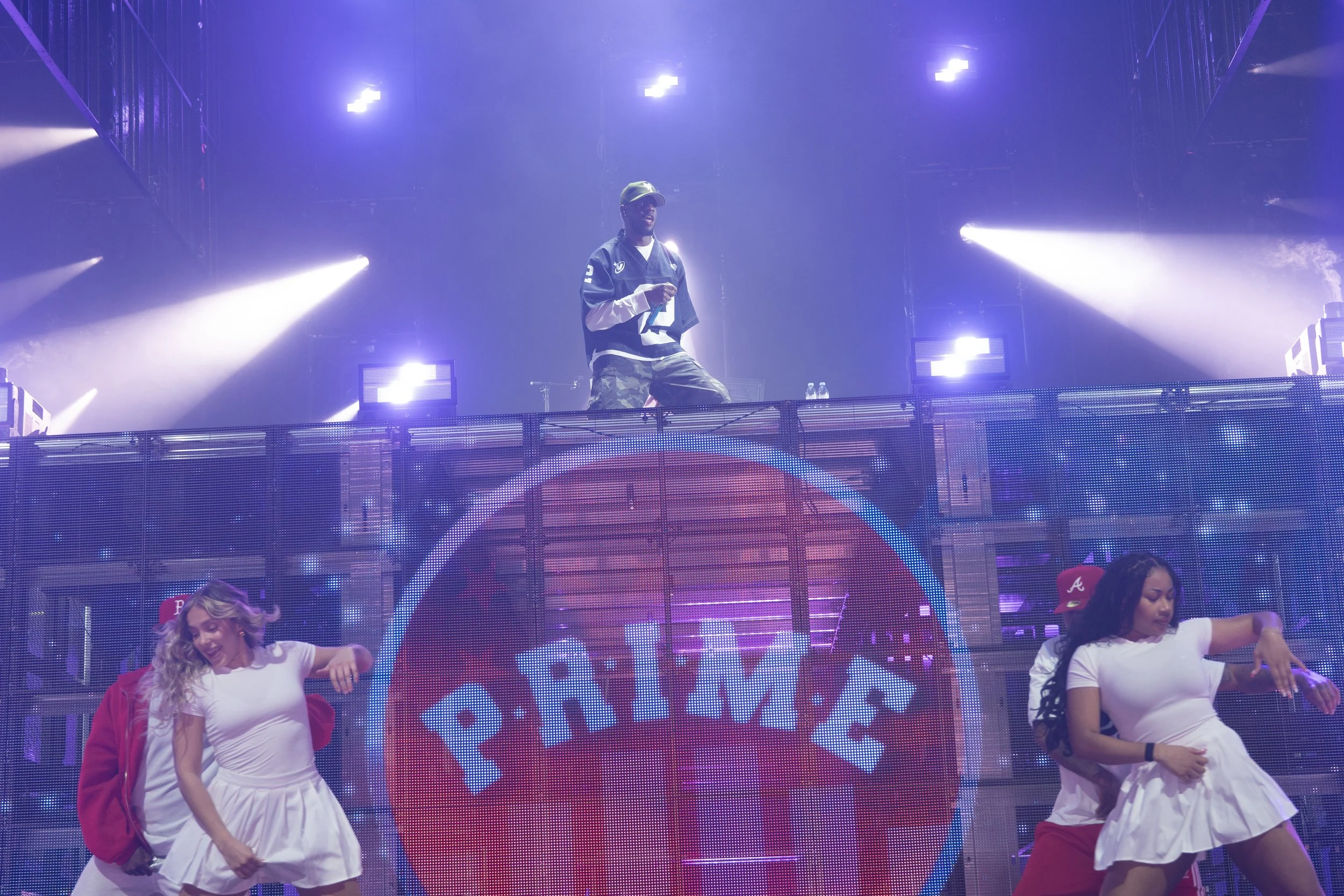 Un DJ joue sur une plateforme avec un panneau indicateur lumineux. En dessous, des danseurs en tenues blanches et rouges dansent dans un environnement lumineux avec des effets de lumière.