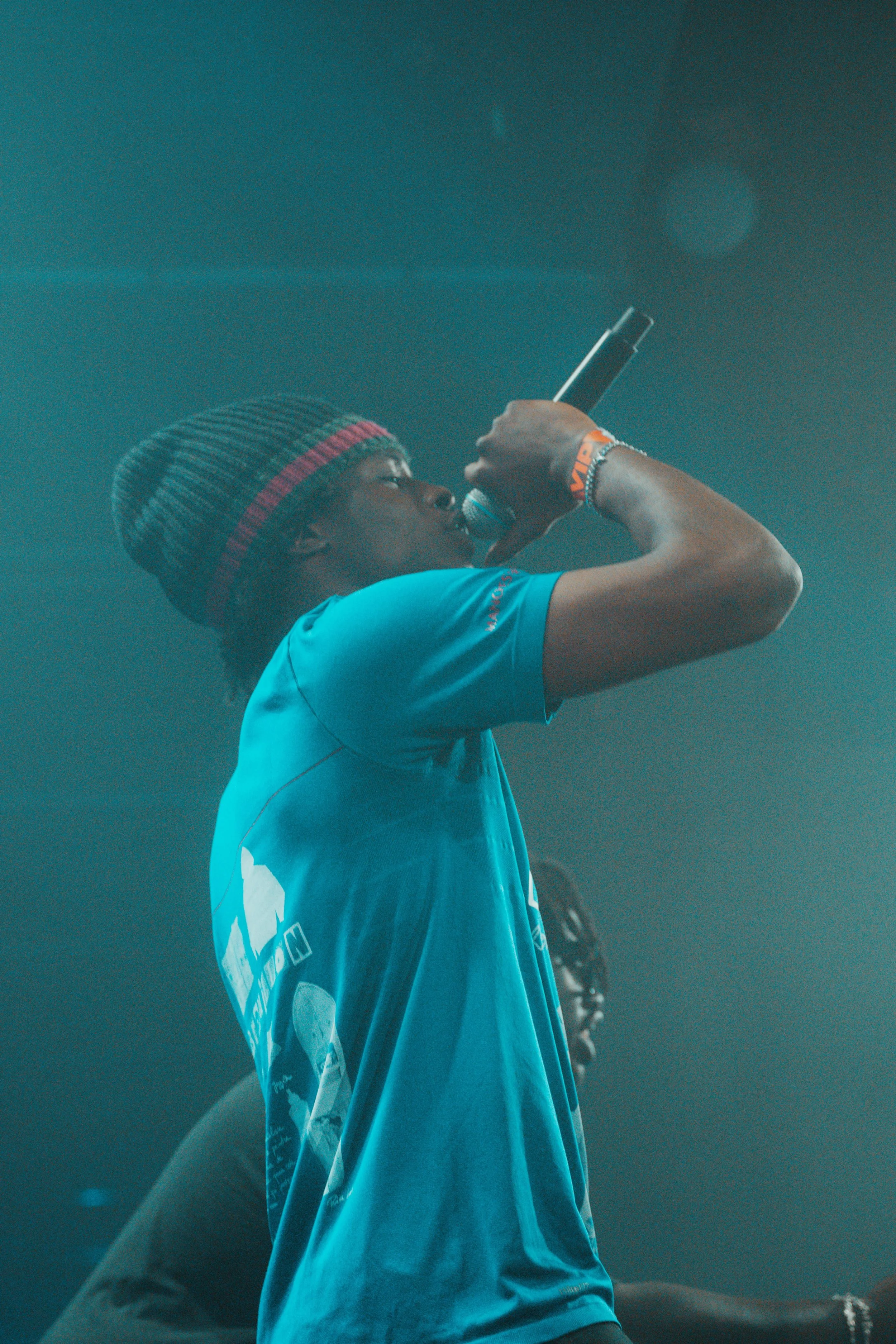 Un jeune homme avec un bonnet à rayures rouges et noires, portant un t-shirt bleu, chante ou parle dans un microphone lors d'une performance ou d'un événement.