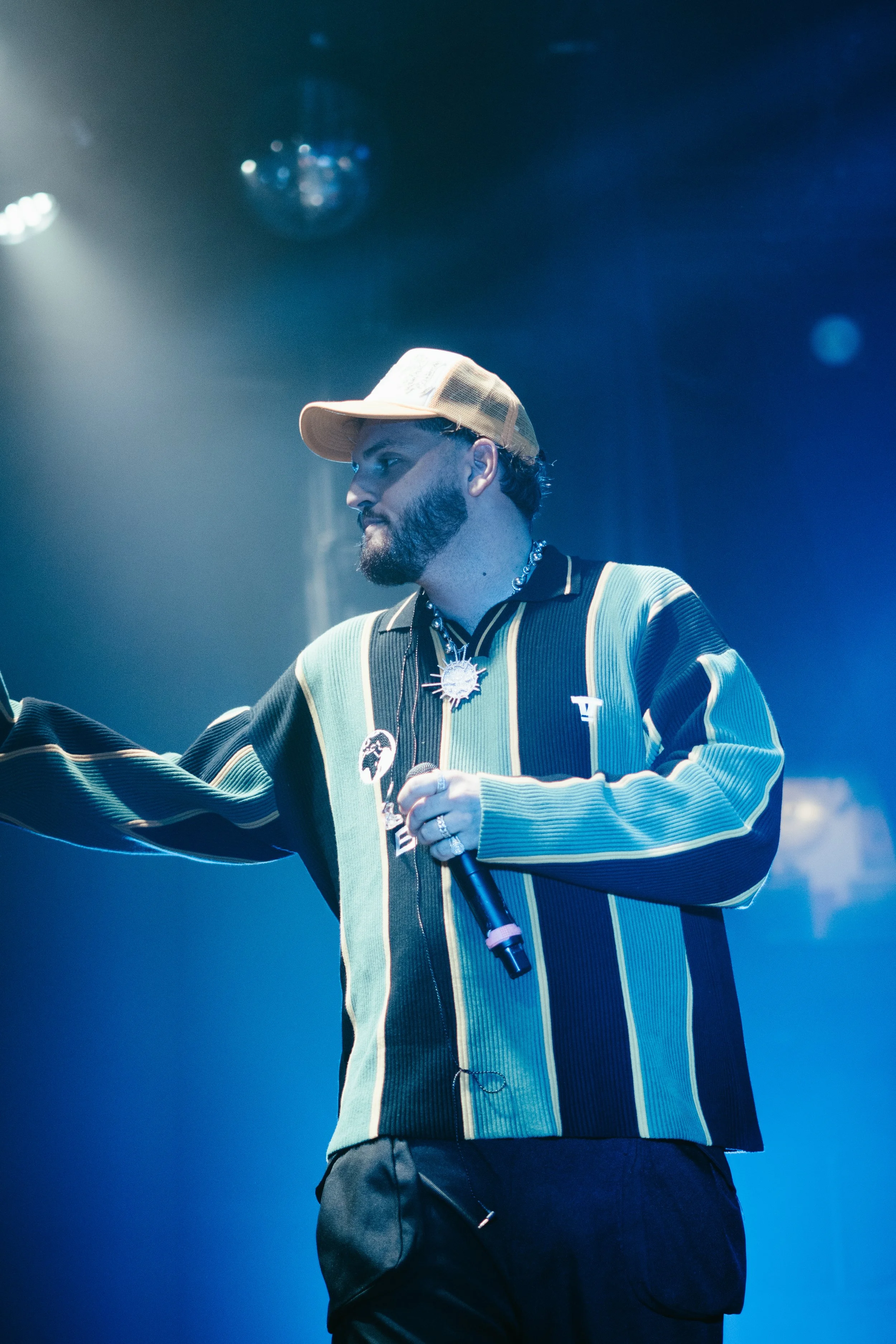 Un homme portant une casquette beige, une veste rayée vert, noir, beige, et blanc, tient un microphone sur scène avec un éclairage bleu.