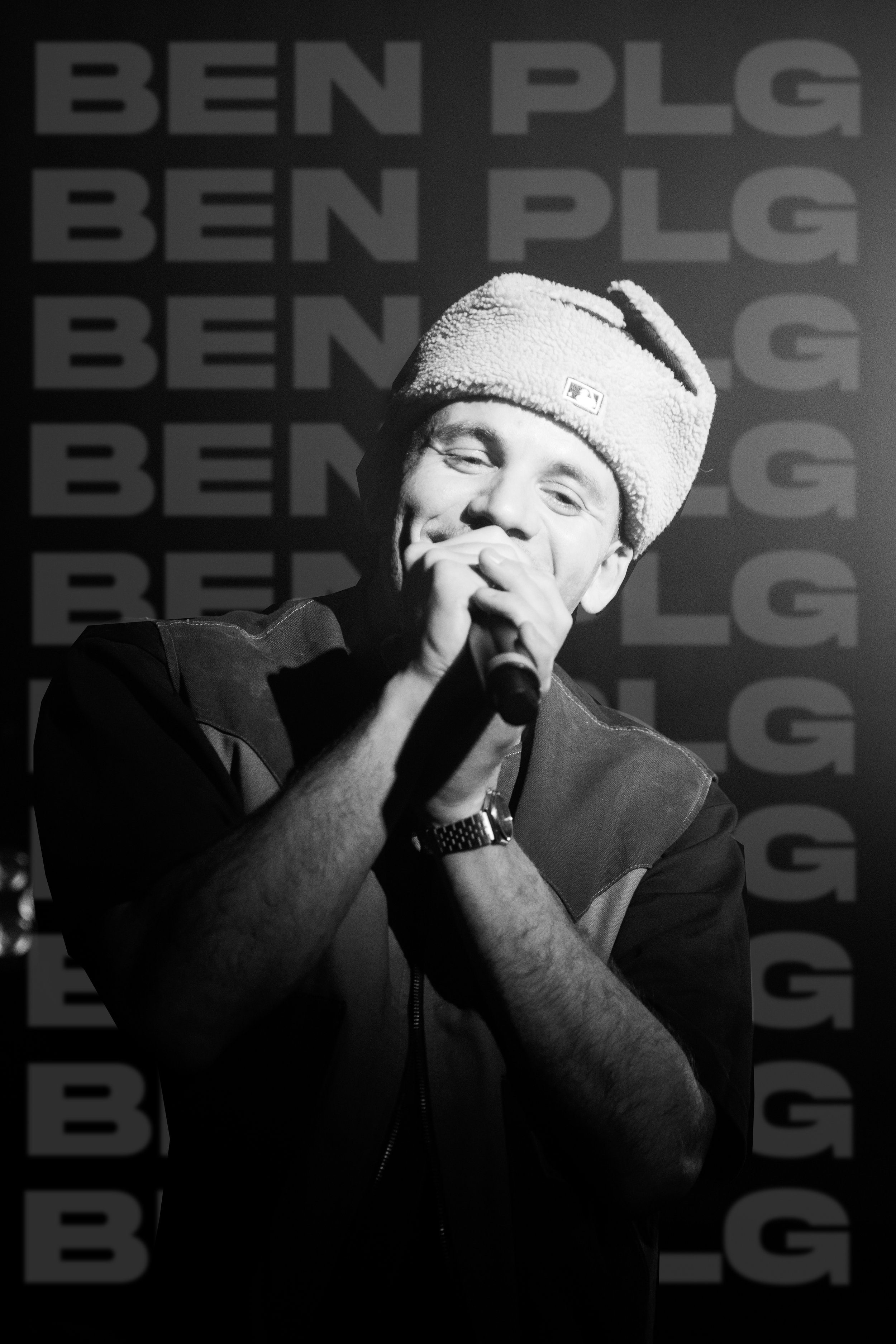 Un jeune homme avec un bonnet en peluche, tenant un microphone, souriant, en noir et blanc, avec un arrière-plan répétant les mots 'BEN PLG'.
