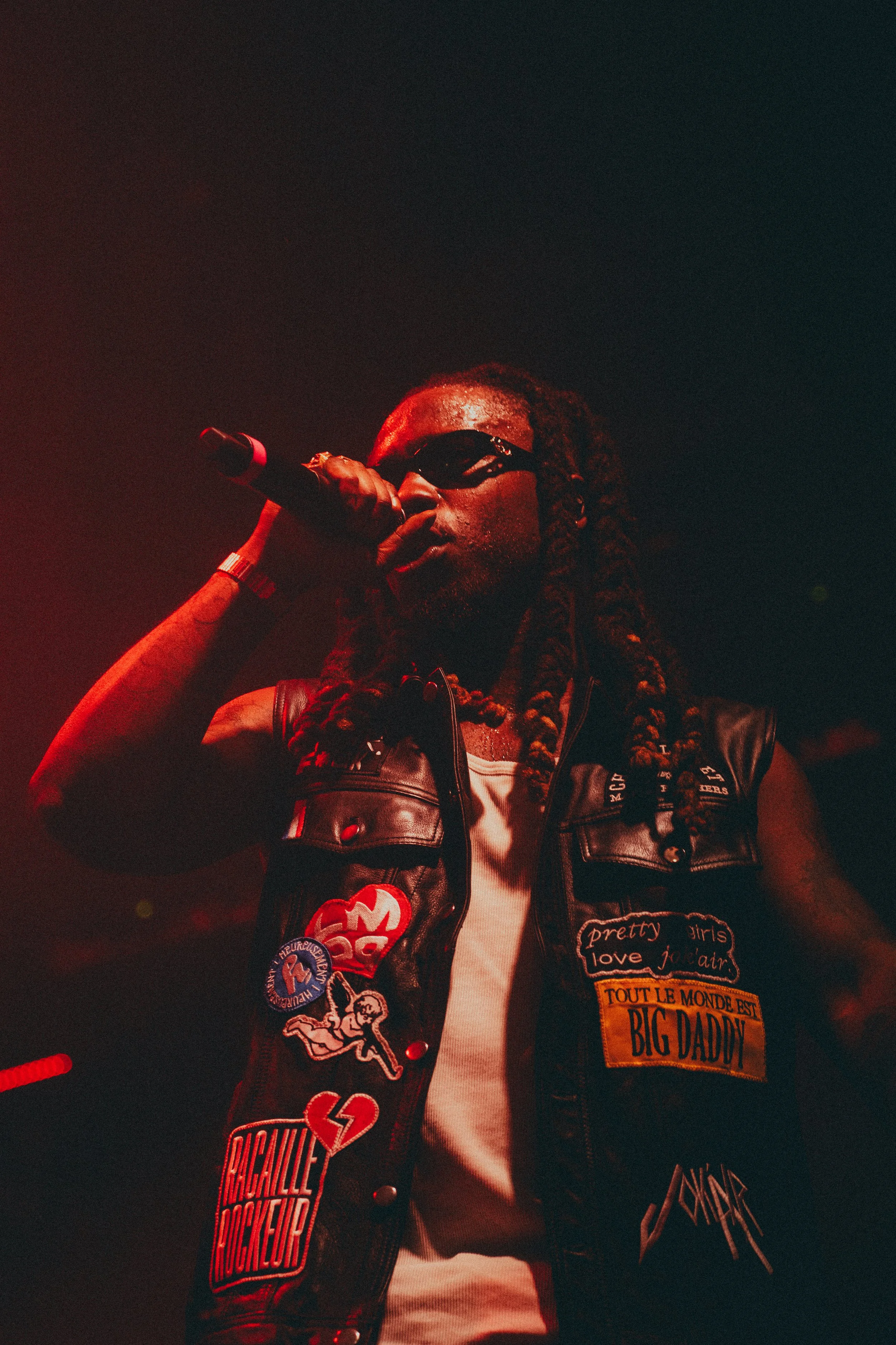Un artiste de rap portant des lunettes noires et une veste en cuir avec des patchs, tenant un micro sur scène dans une ambiance sombre avec un éclairage rouge.