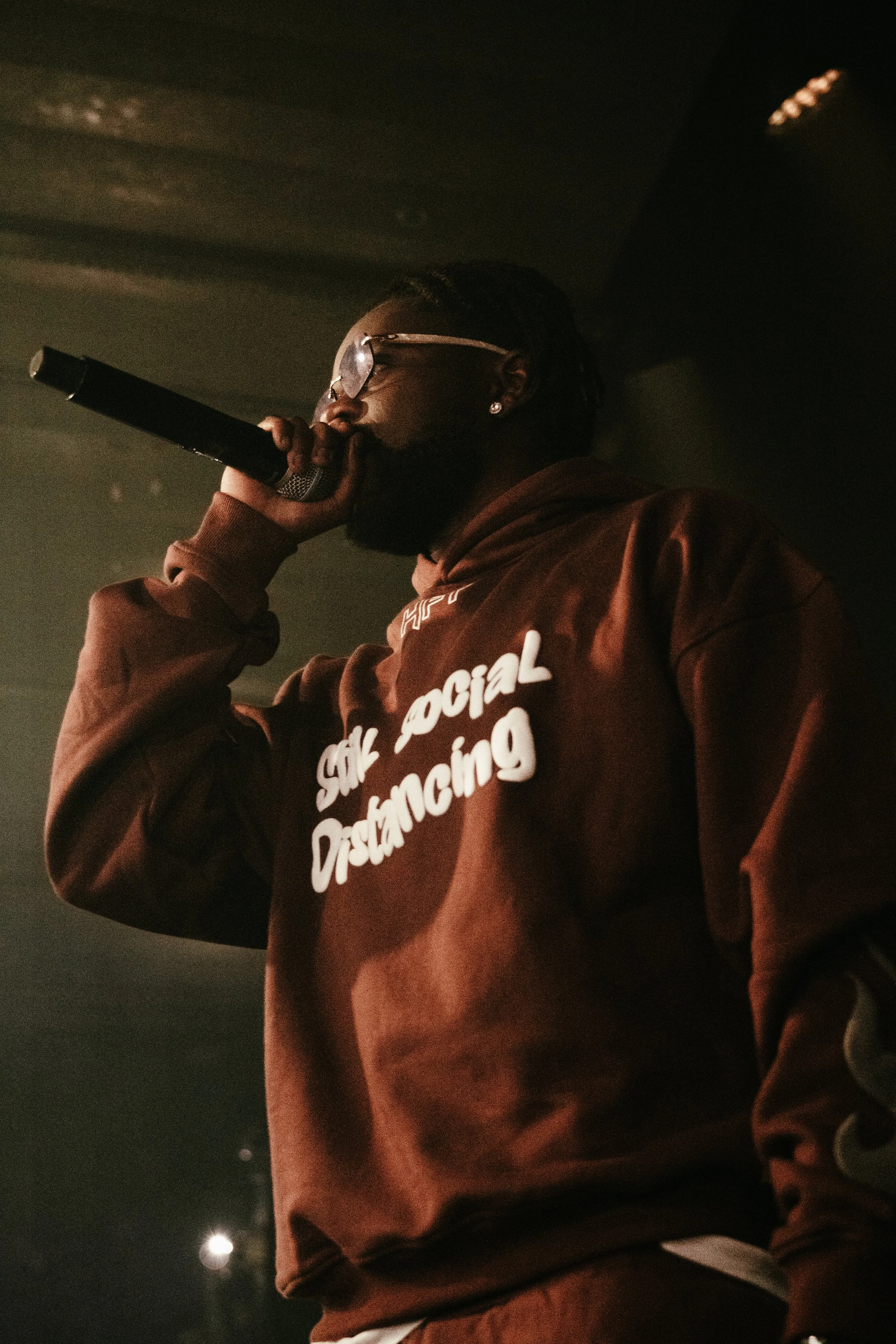 Un homme noir portant un sweat marron avec l'inscription 'SIX SOCIAL Disdancing', portant des lunettes, tenant un microphone, sur scène