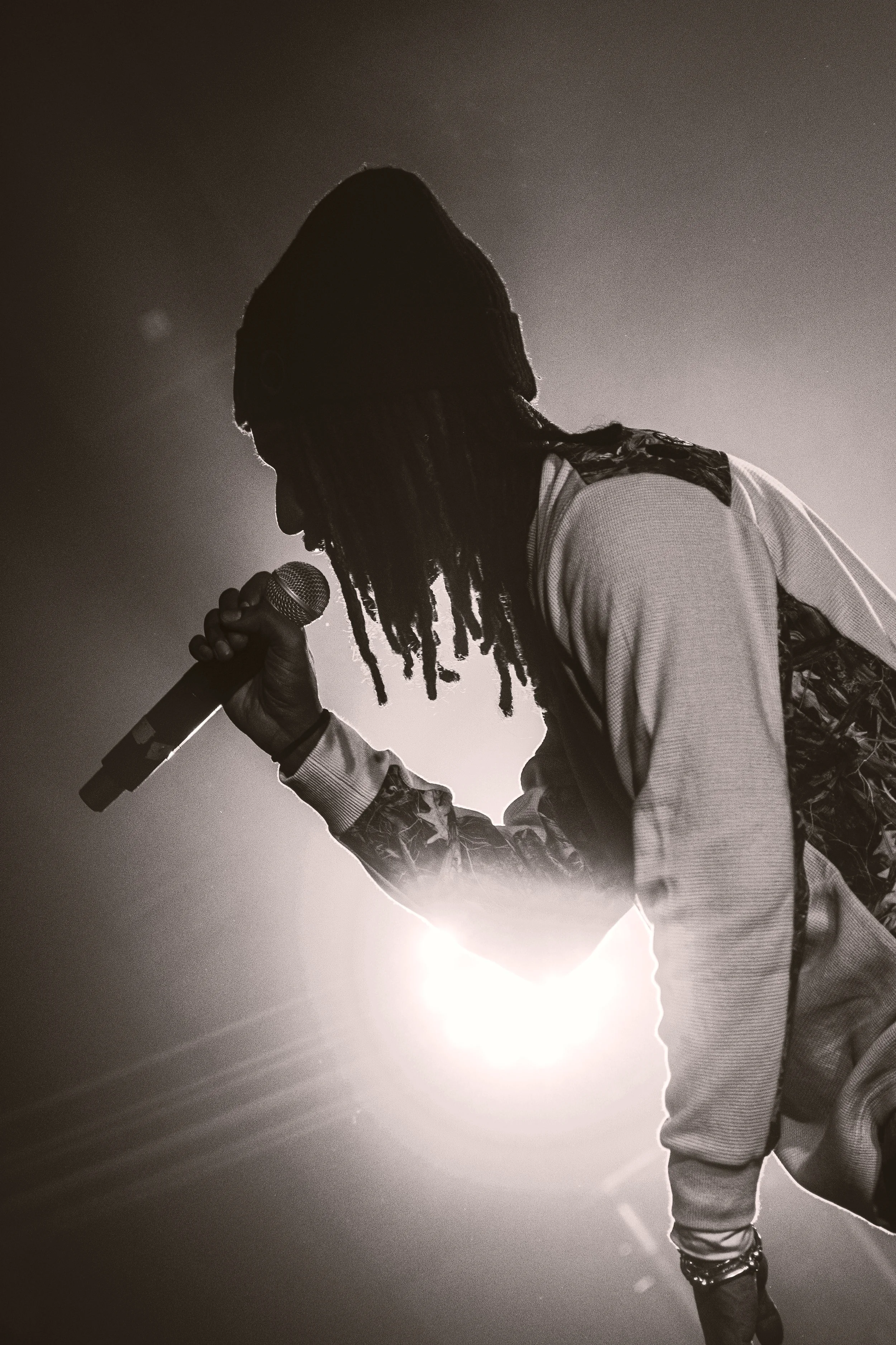 Une personne avec des dreadlocks, en silhouette, tenant un microphone, éclairée par une lumière forte derrière elle.