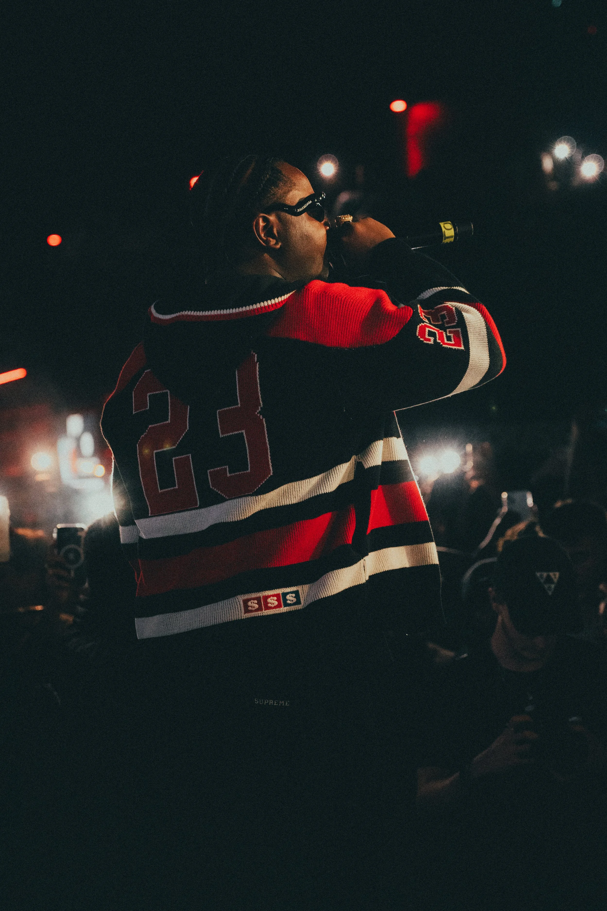 Une personne portant un maillot de hockey noir, rouge et blanc avec le numéro 23, chante ou parle dans un microphone dans un environnement nocturne, entourée d'autres personnes.