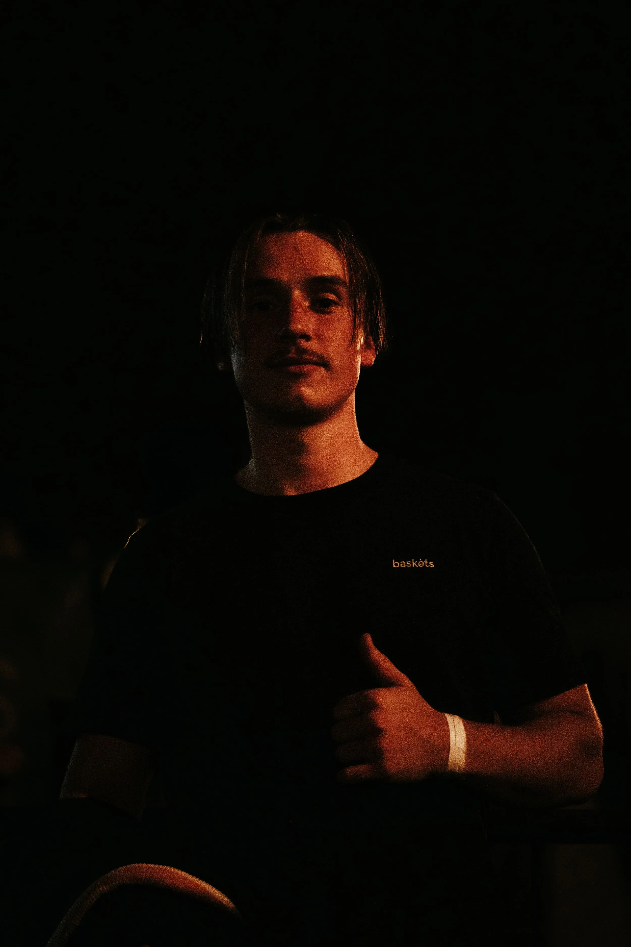Jeune homme avec des cheveux mi-longs, portant un t-shirt noir avec le mot 'baskêts' en petit texte orange, tenant un pouce levé, dans un environnement sombre. 