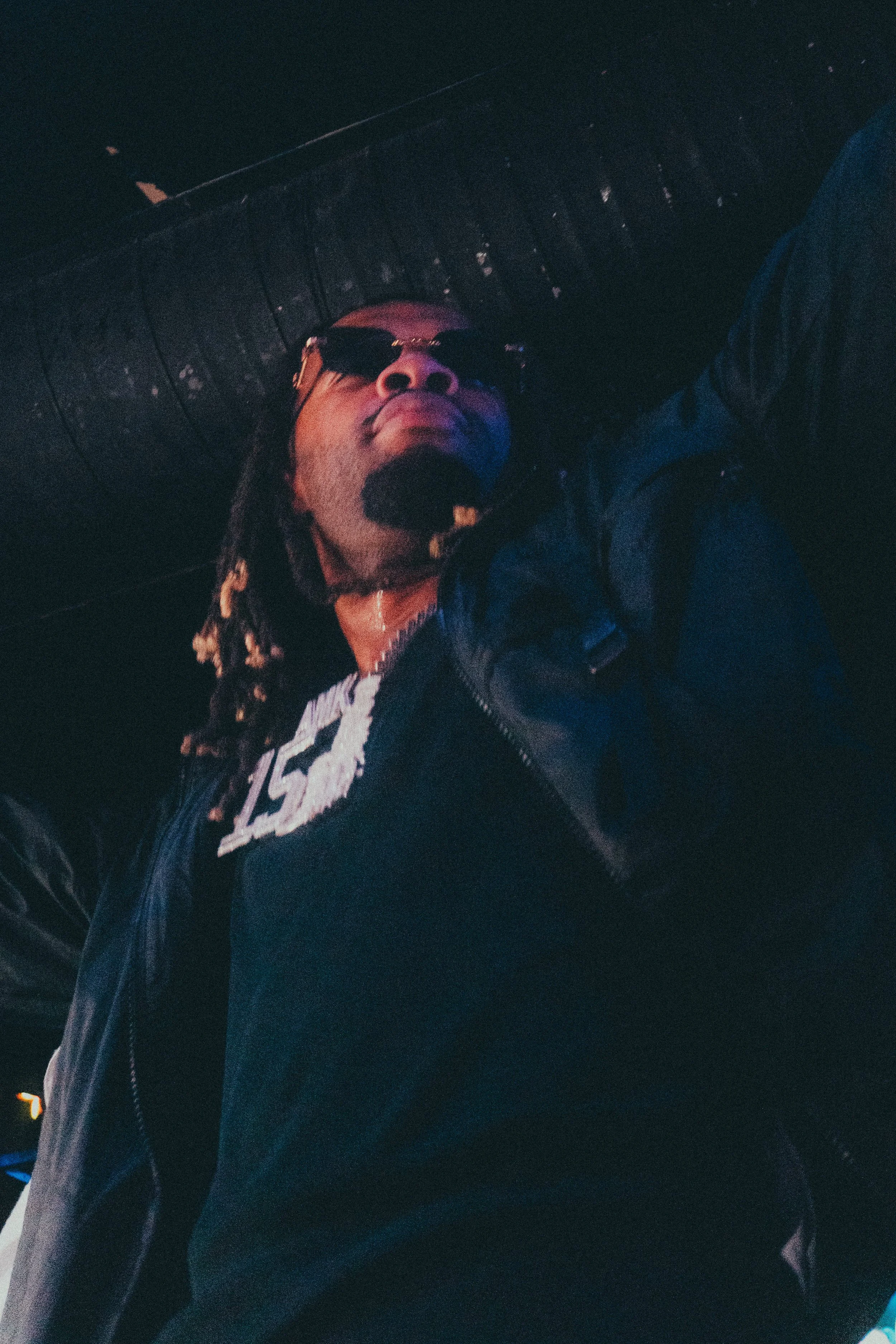 Un homme portant des lunettes de soleil, une veste en cuir, haute dans un environnement sombre avec un plafond en bois, éclairé par des lumières faibles.