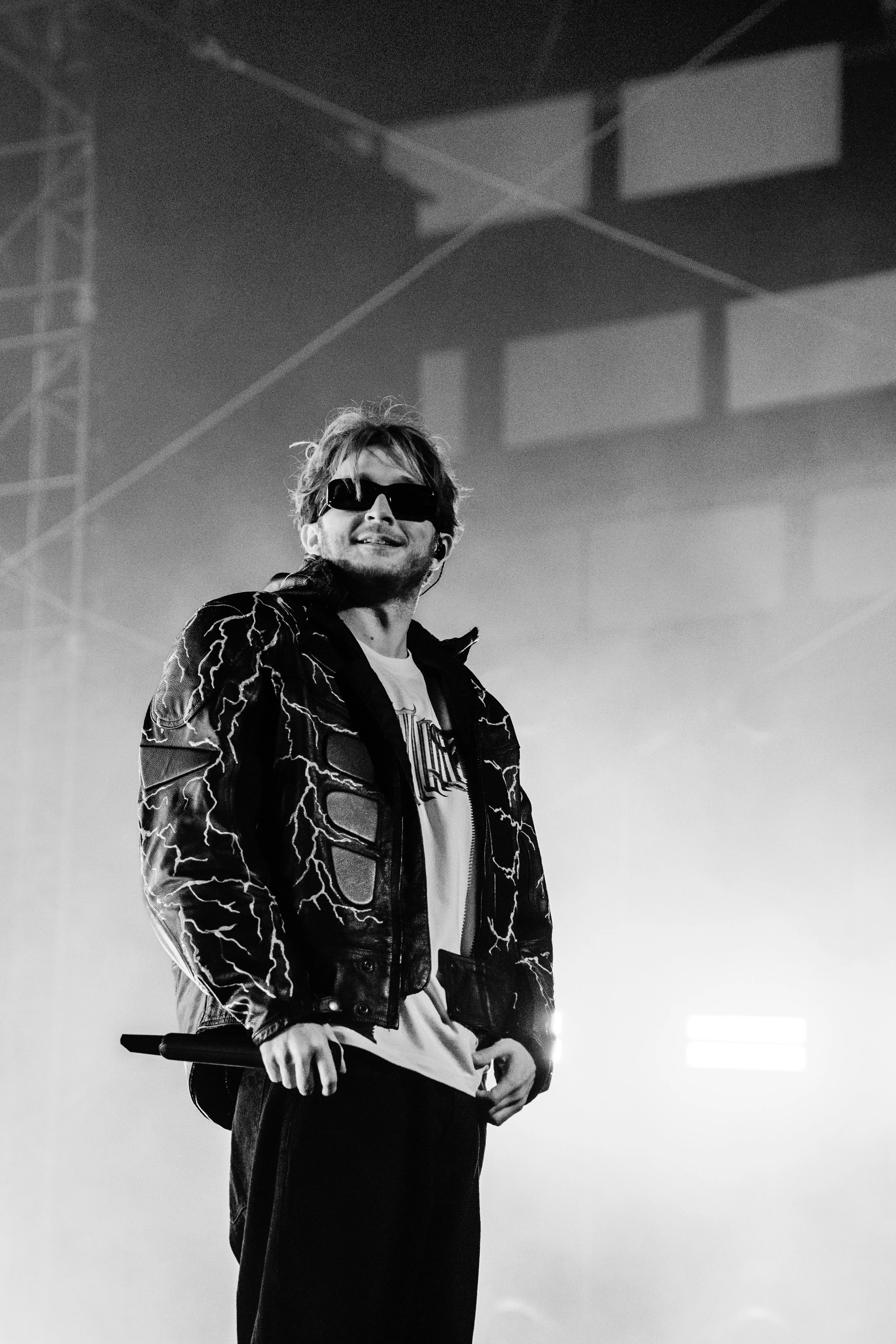Un homme souriant portant des lunettes noires et une veste en cuir avec des motifs électriques, tenant un micro sur scène dans une ambiance sombre.