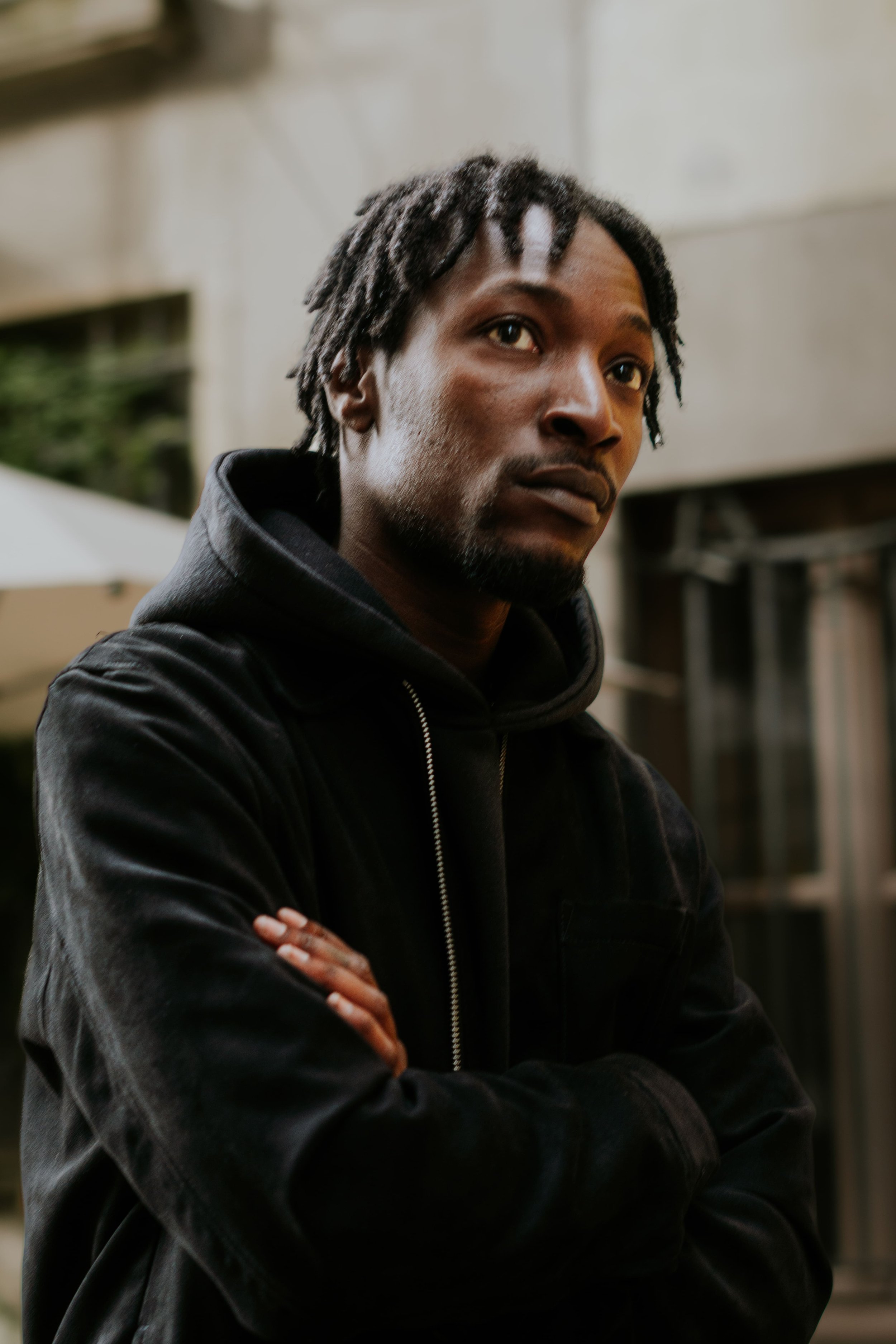 Jeune homme noir avec dreadlocks, portant un sweat à capuche noir, croisant les bras, dans un environnement urbain.