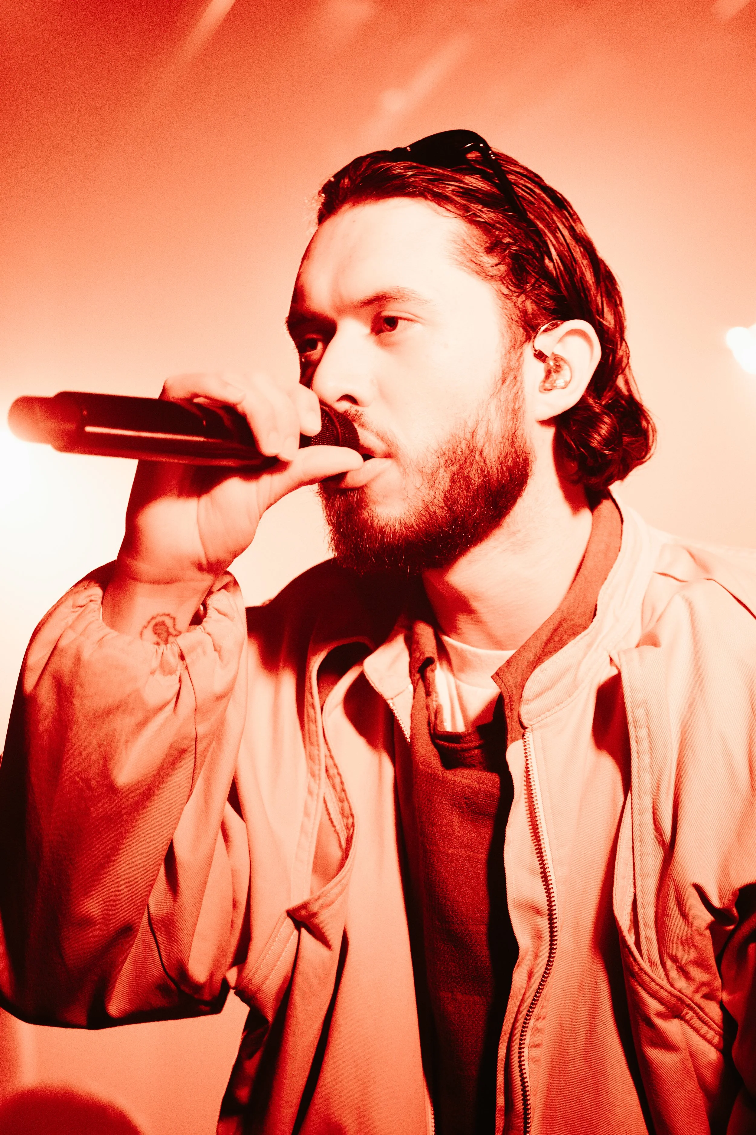 Un homme avec des cheveux longs et une barbe, portant une veste beige, tient un microphone et chante lors d'une performance, un éclairage rougeâtre en arrière-plan.