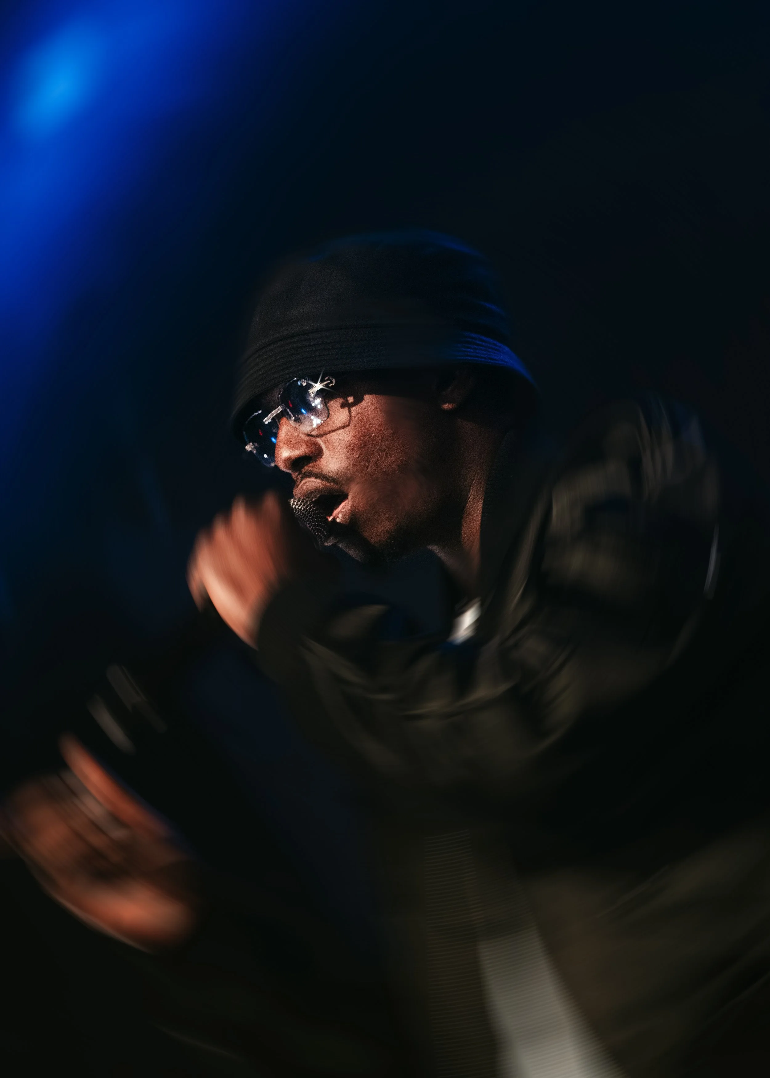 Un homme chantant ou parlant dans un microphone, portant des lunettes de soleil et un bonnet, dans un environnement sombre avec un éclairage bleu.